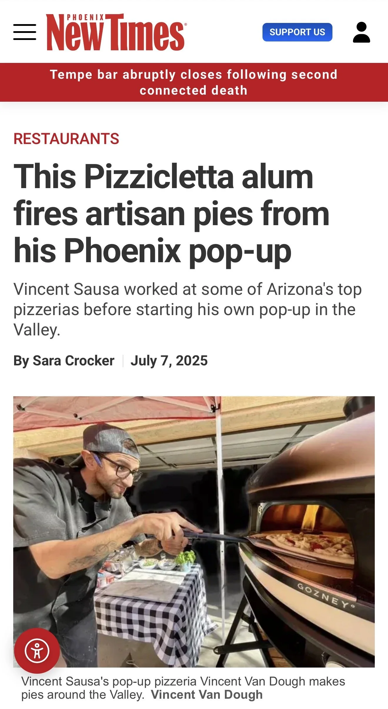 A man wearing glasses, a black cap, and a dark chef's coat is placing pizza pies into an outdoor pizza oven with a Gozney logo. There is a checkered tablecloth on a table nearby and a people tent above.