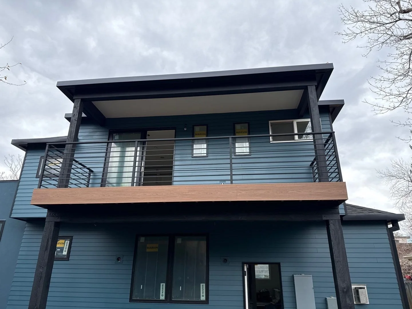 Two-story modern house with blue siding, black accents, a large front balcony with metal railing, and a flat roof with extended overhangs. Bare trees are visible on the sides, and the sky is cloudy.
