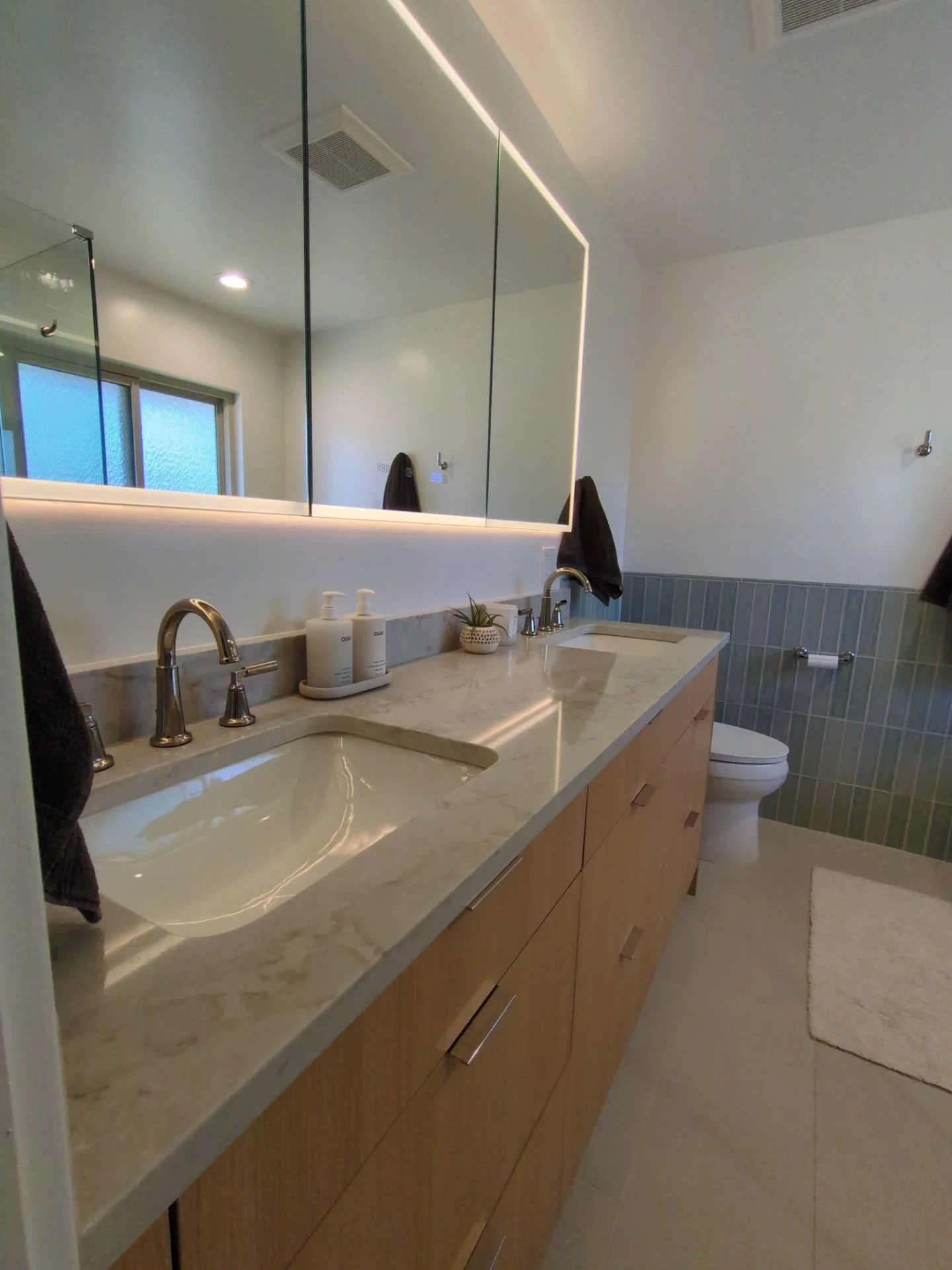 Modern bathroom with a marble countertop, undermount sink, wooden cabinetry, large mirror, toiletries, a potted plant, towel hooks, and a toilet with light green tiled wall behind it.