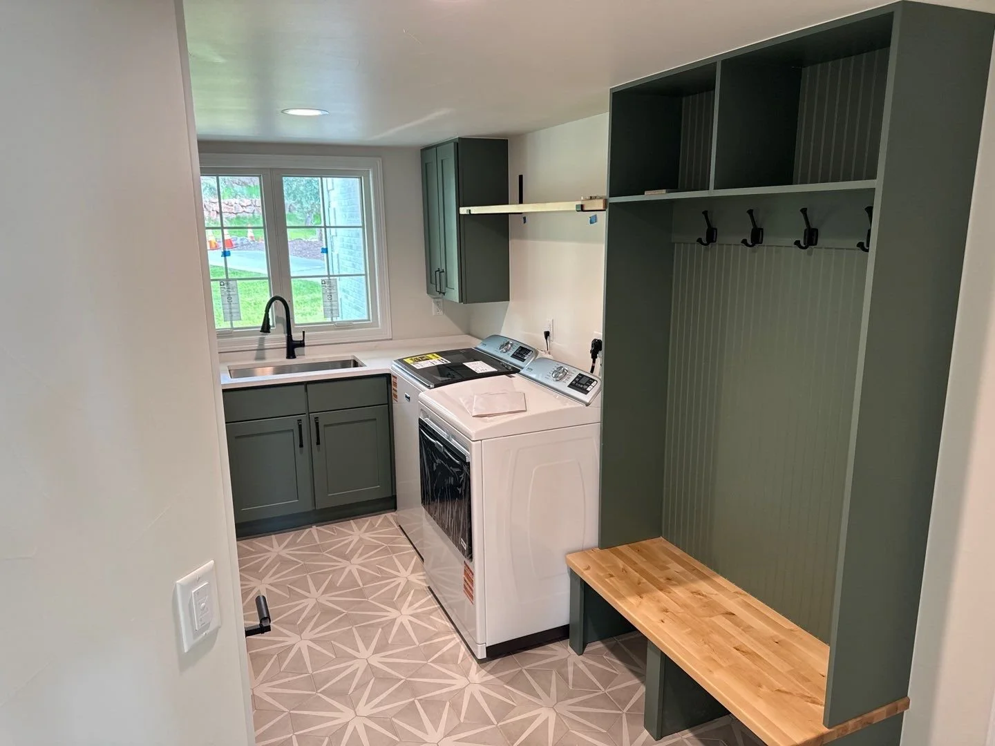 A laundry room with green cabinets, a white washer and dryer, a black sink faucet, a window with a backyard view, a wooden bench, and a green wall with hooks.