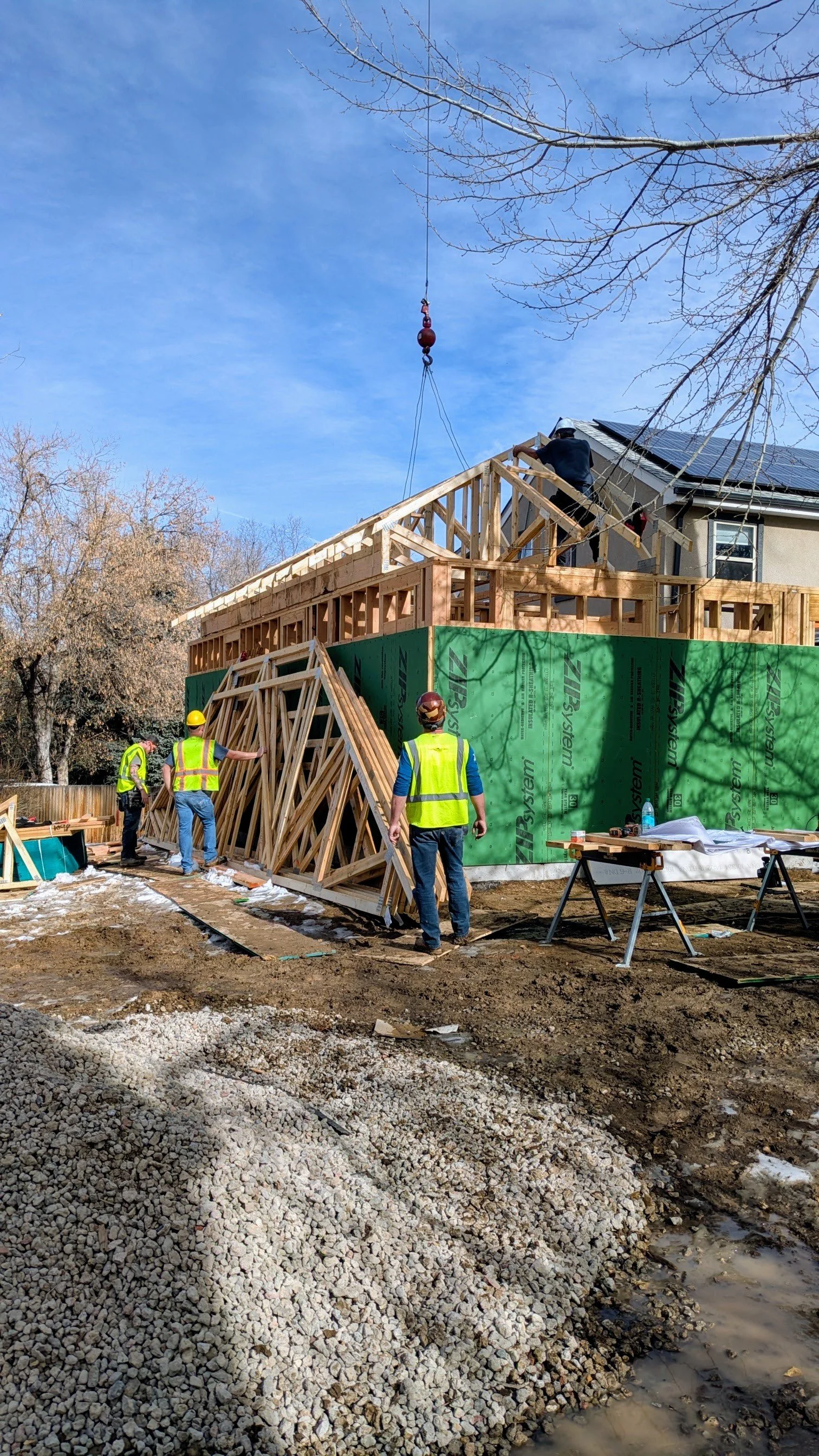 Construction workers building a residential house, with some workers guiding the framing and others working on the roof, using tools and wearing safety gear, with a crane in the background lifting materials.