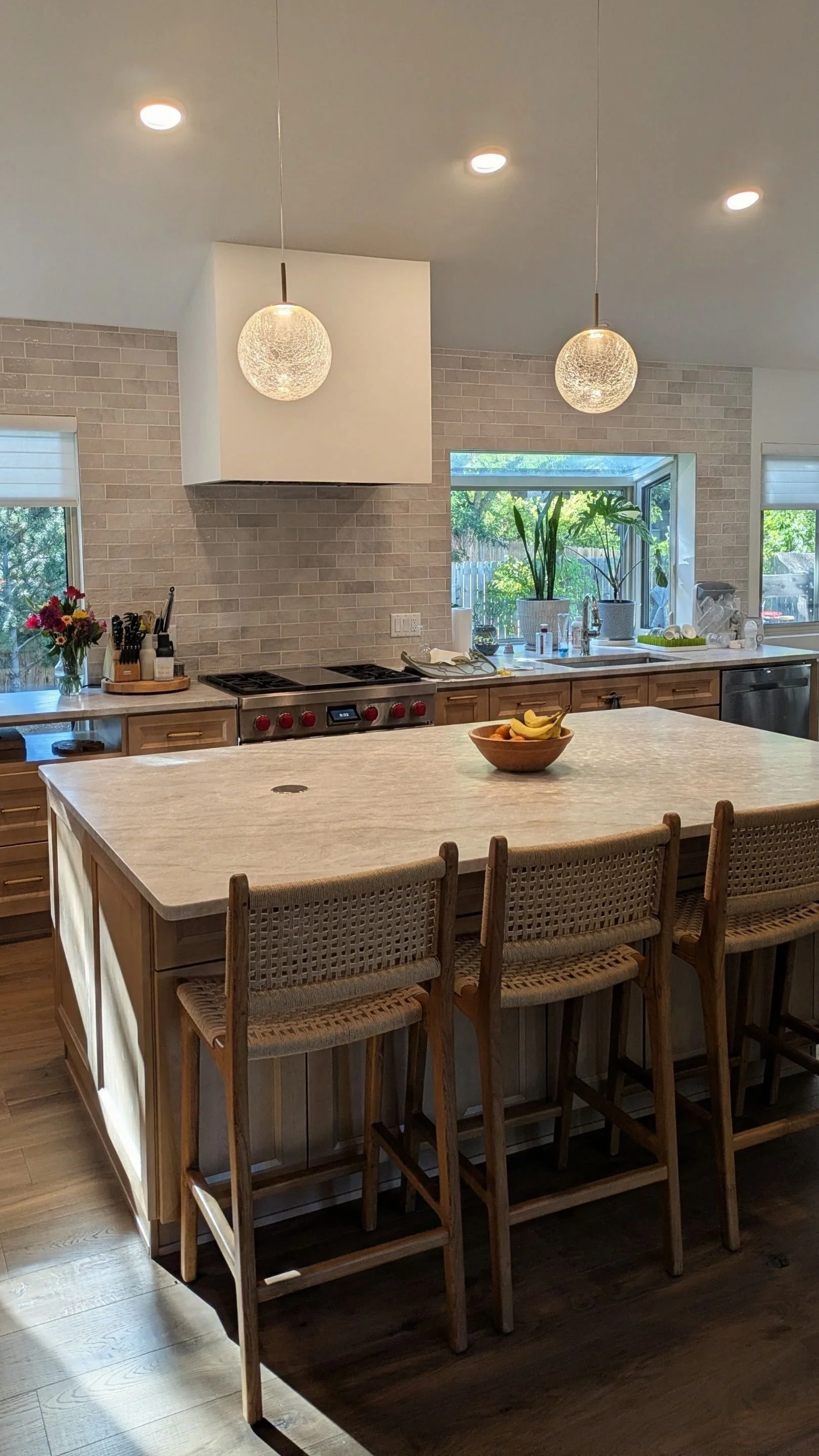 A modern kitchen with a large island, wooden cabinets, a stove, and a window with greenery outside. The island has a bowl of bananas, and there are two pendant lights hanging from the ceiling.