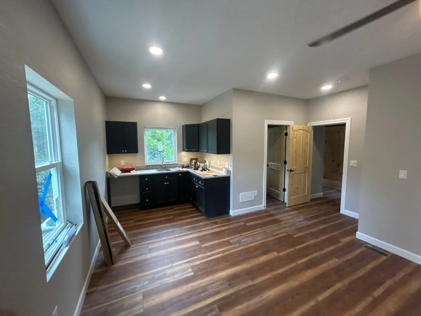 Empty kitchen with gray walls, black cabinets, wood floors, and a window with a view of trees; some tools and supplies on the counter.