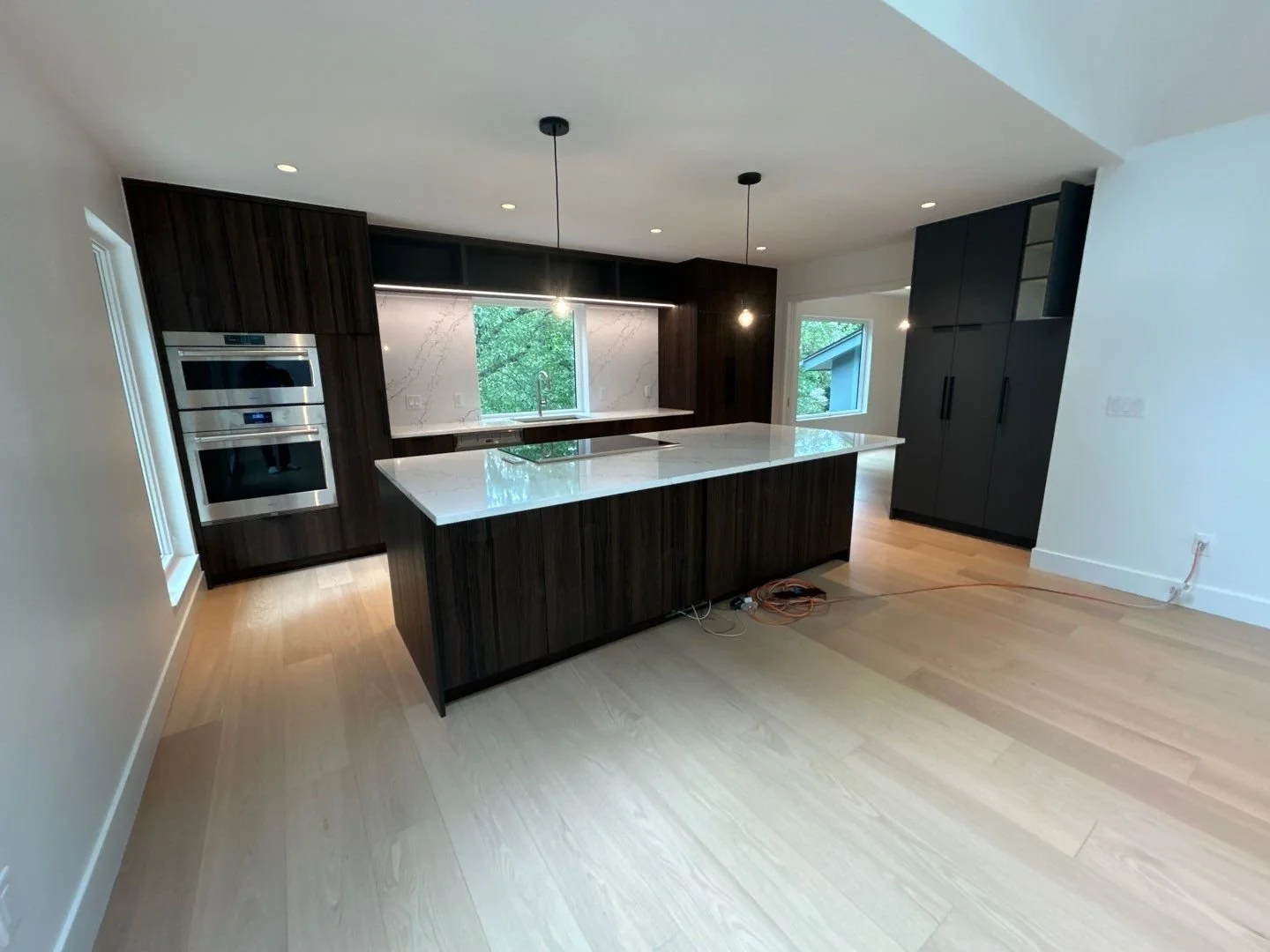 Modern kitchen with dark wooden cabinets, white marble countertops, built-in double ovens, large island, a window above the sink, and light hardwood flooring.