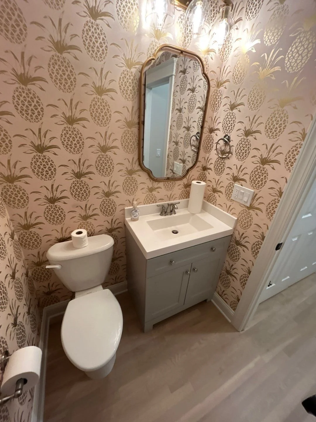 Bathroom with pineapple-patterned wallpaper, a white toilet with a roll of toilet paper on top, a small vanity with a sink, mirror, and light fixtures above.