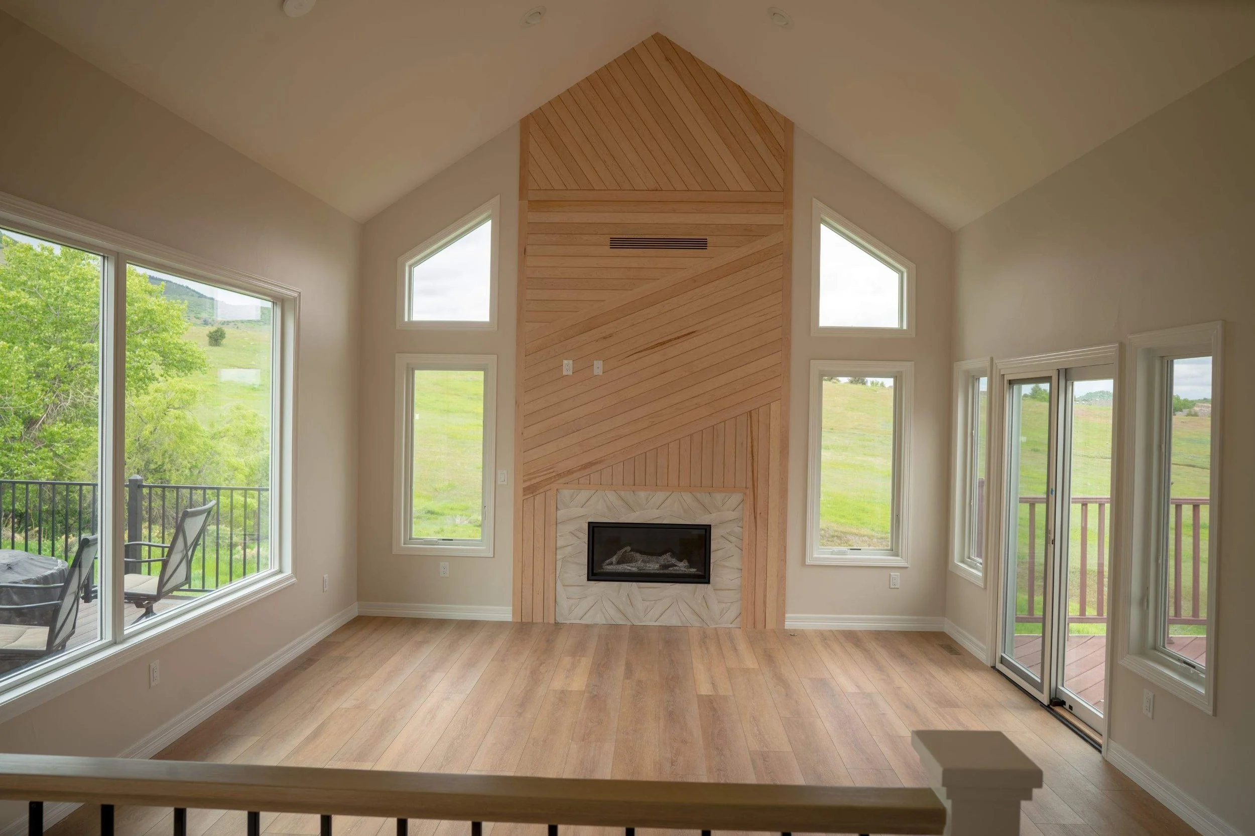 Interior view of a living room with a central fireplace surrounded by a wooden mantel, large windows showing a green landscape outside, hardwood floors, and a vaulted ceiling.