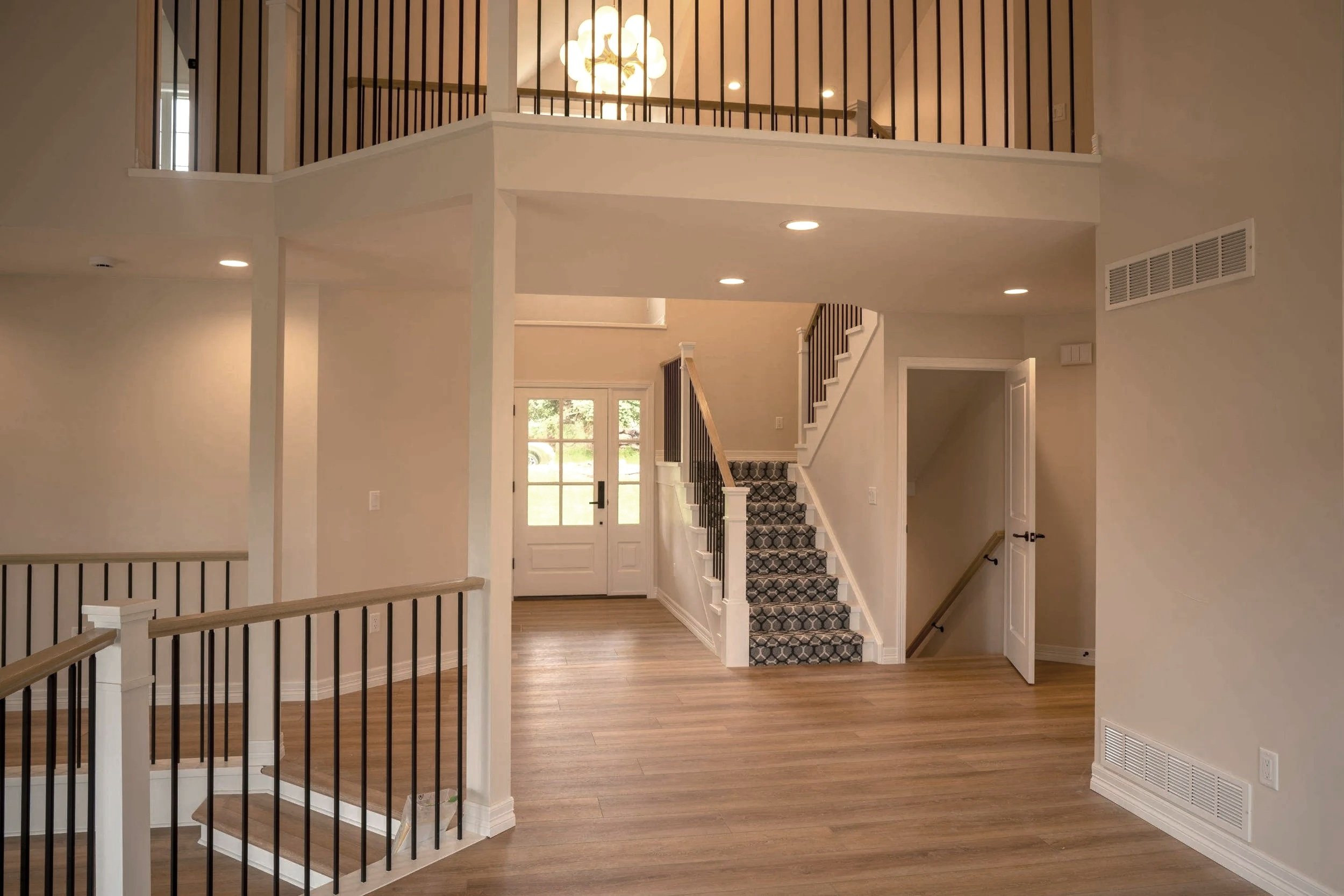 Interior view of a modern home entrance with wooden flooring, staircase with patterned carpet, and a hallway leading to a door with glass panels. The ceiling has recessed lights, and there is a railing for a second-floor balcony.