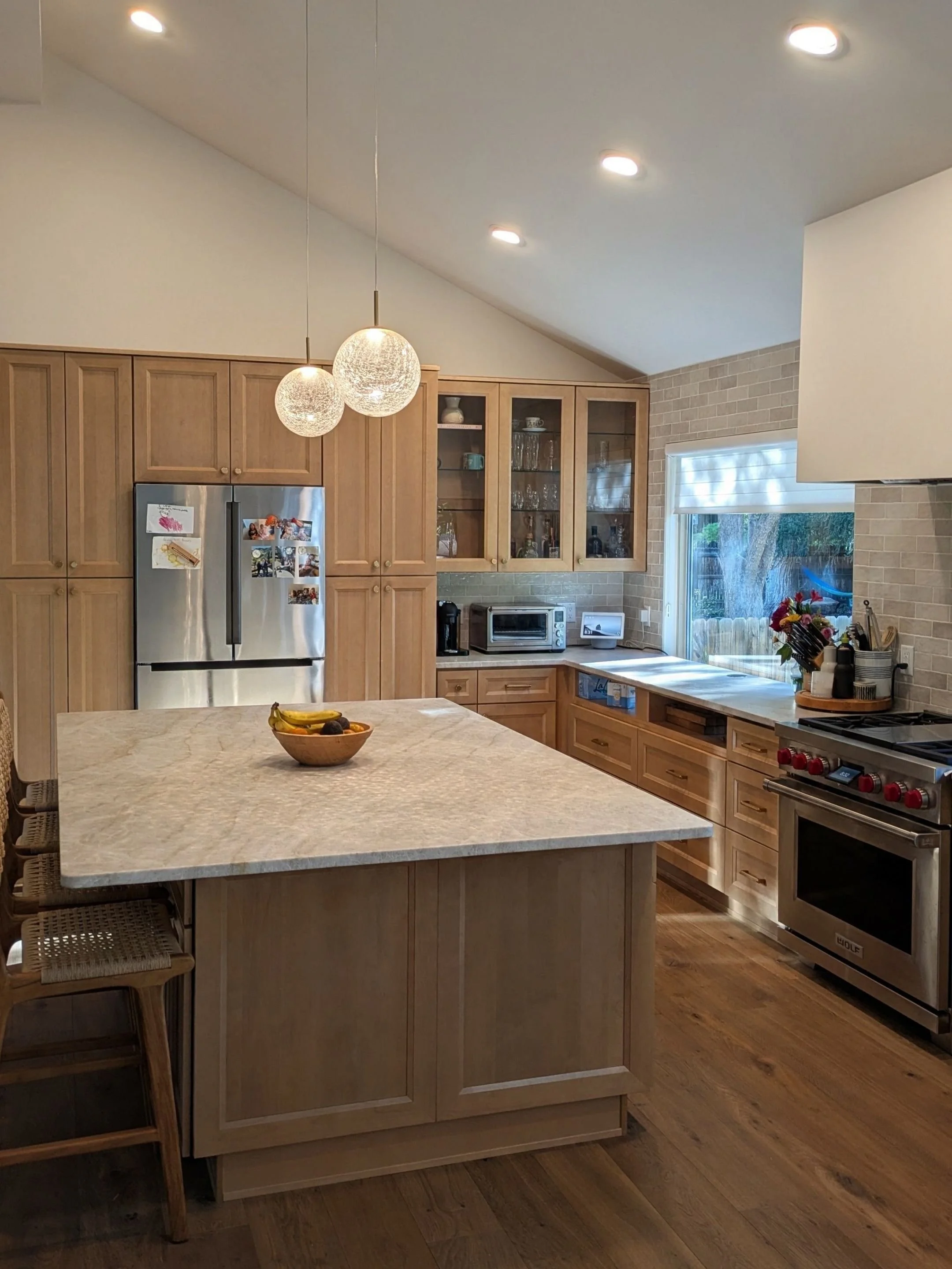 A modern kitchen with wooden cabinets, a central island with a marble countertop, a stainless steel refrigerator with family photos, a microwave, a toaster, a stove, and a window with a view of trees outside.