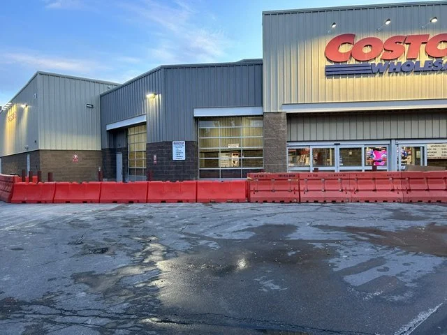 Costco Wholesale store with a gray exterior, red barrier in the foreground, and a wet parking lot.