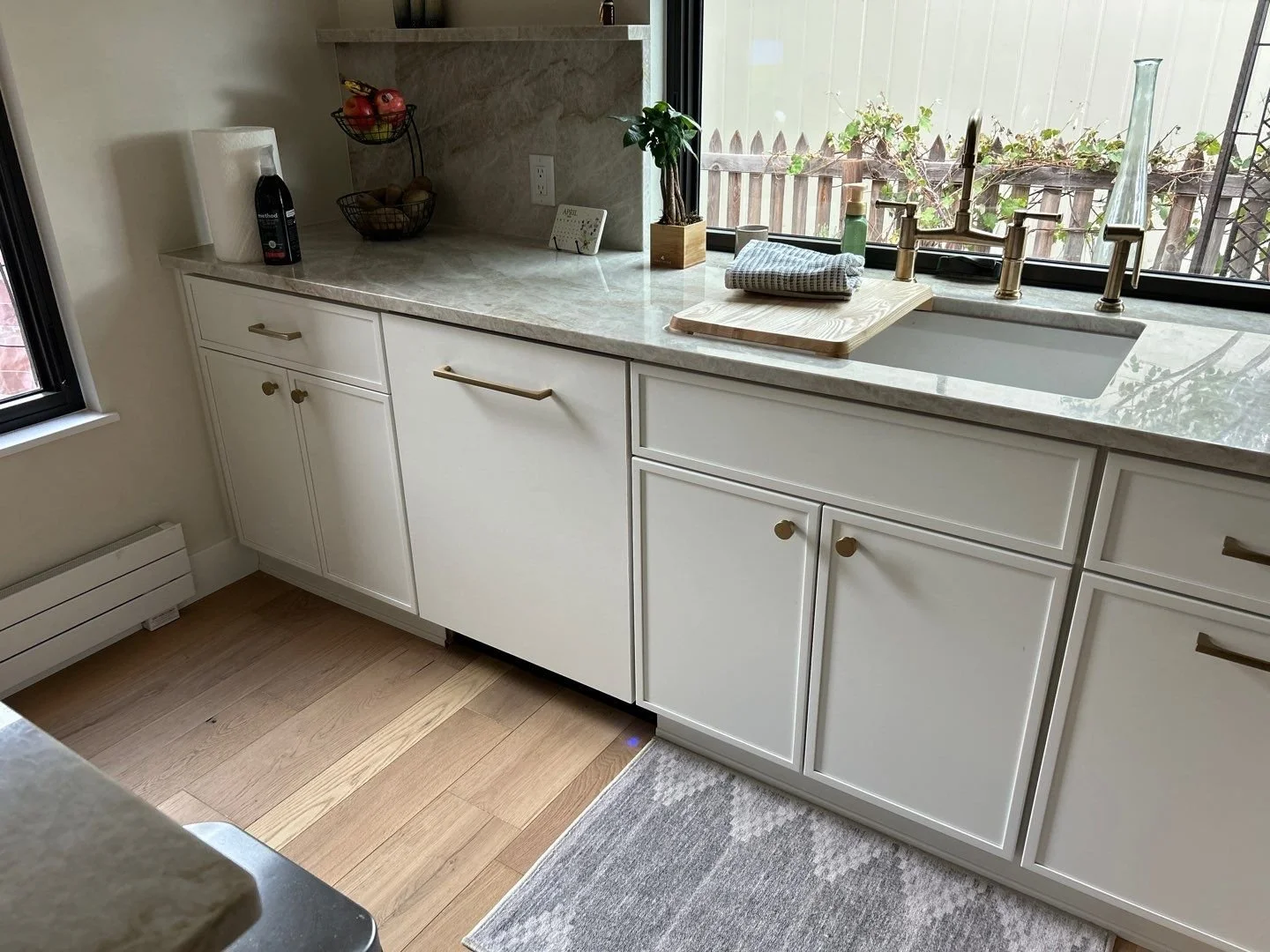 Kitchen sink area with white cabinets, a marble countertop, a brass faucet, a window above the sink, and decorative items including a small plant, a soap dispenser, a dish towel, and a cutting board with a cloth on it.