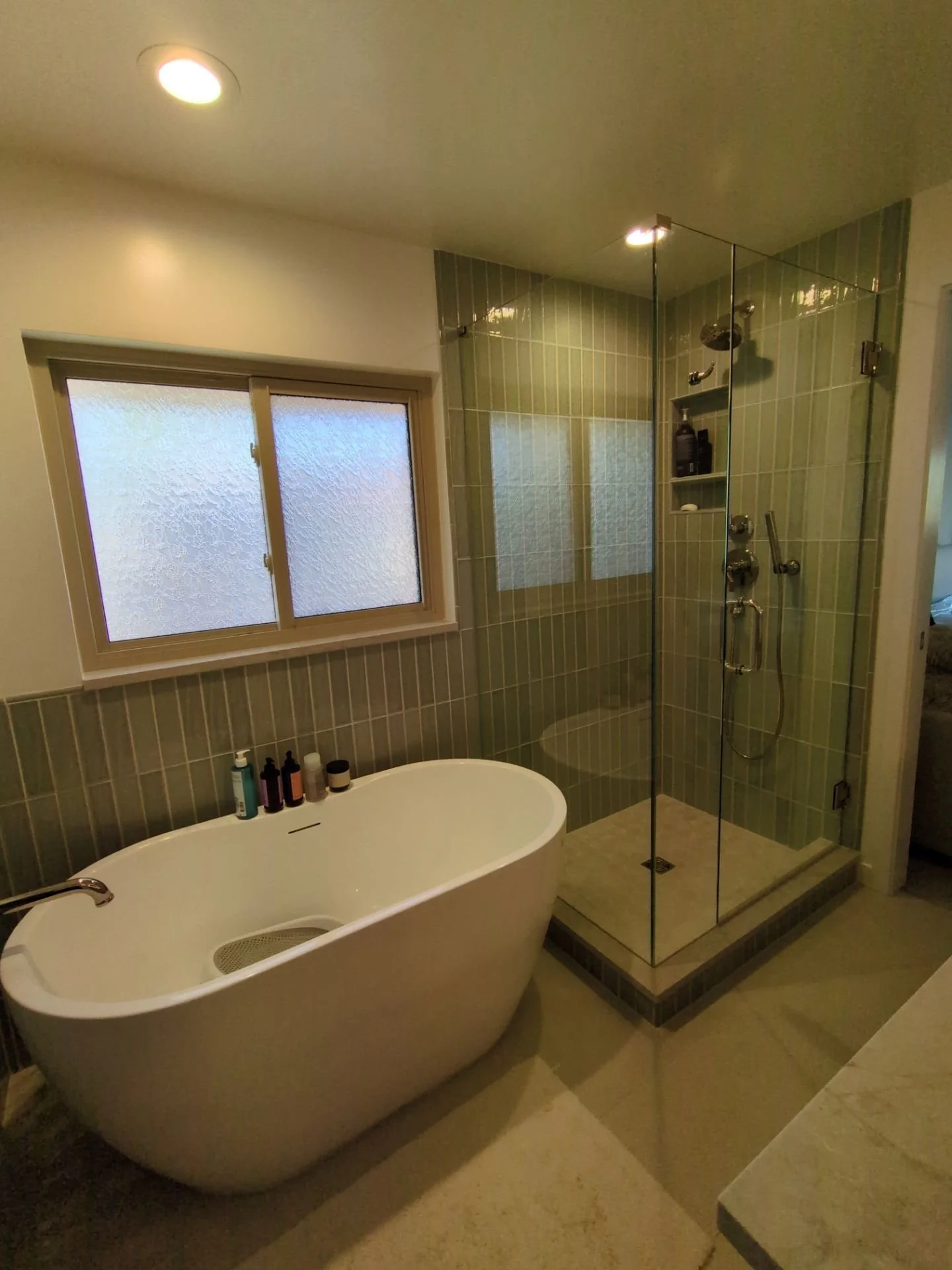 Bathroom with a freestanding bathtub, a window with frosted glass, and a glass-enclosed shower with green tiles.