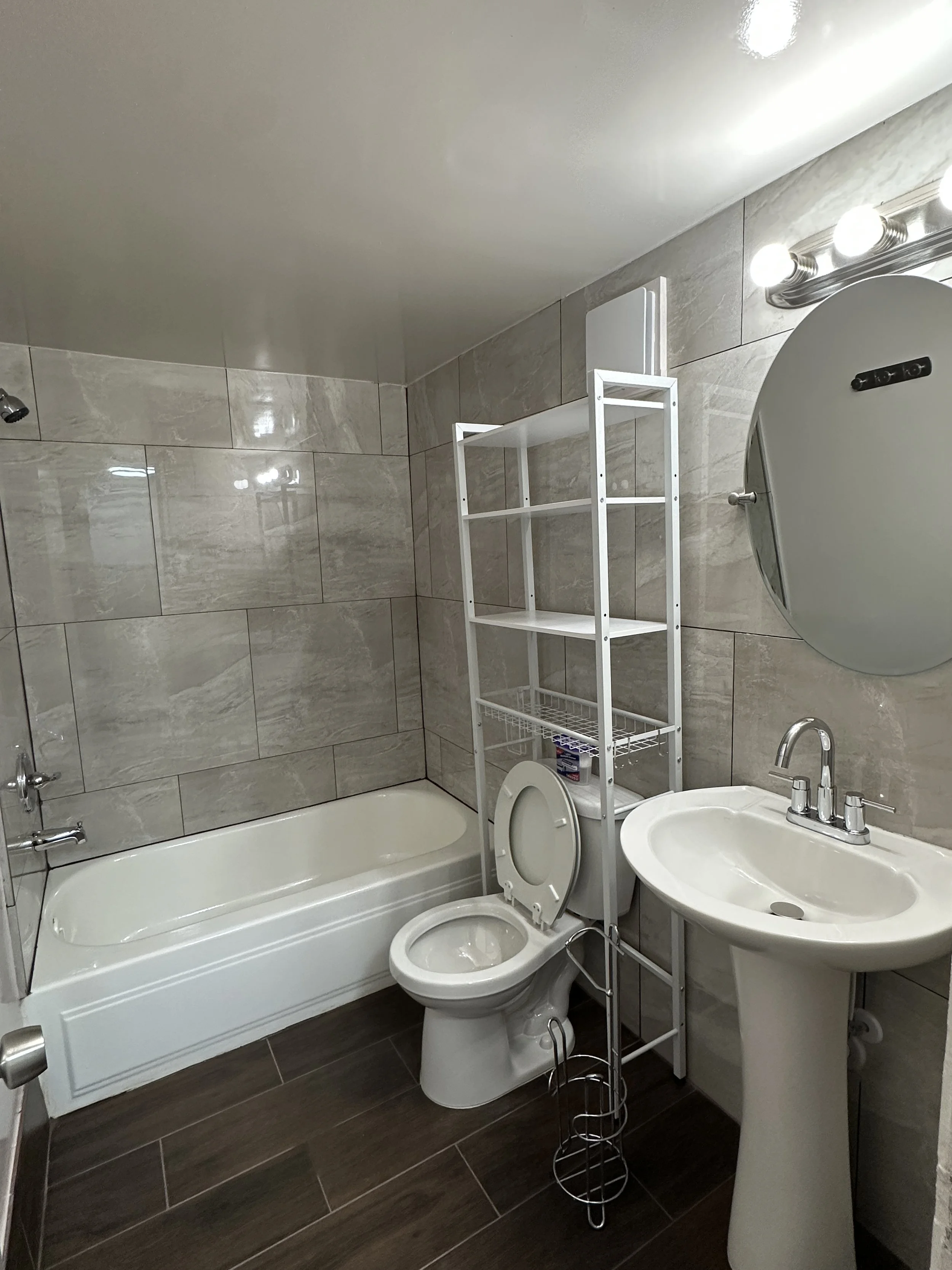 A small bathroom with beige tiled walls, a bathtub, a toilet, a white shelf, a sink, and a mirror with three light bulbs.