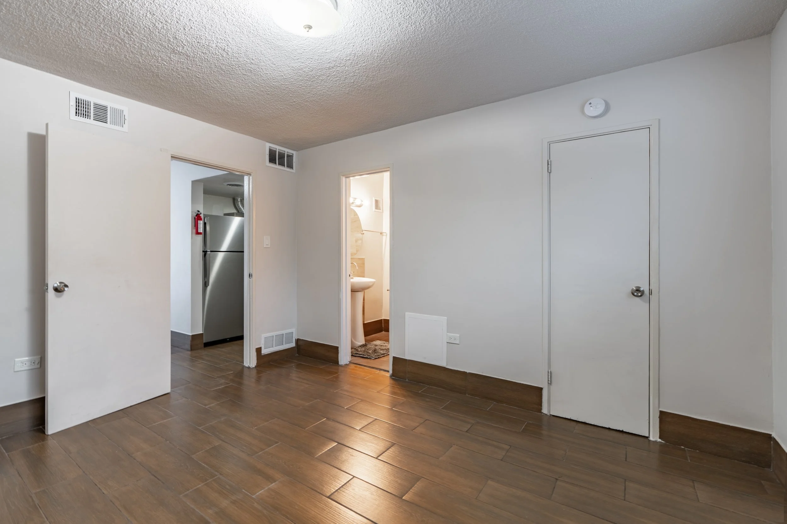 Empty room with white walls, brown wooden floor, and multiple doors, including a view into a bathroom with a sink and mirror, ceiling light, wall vents, and a smoke detector.