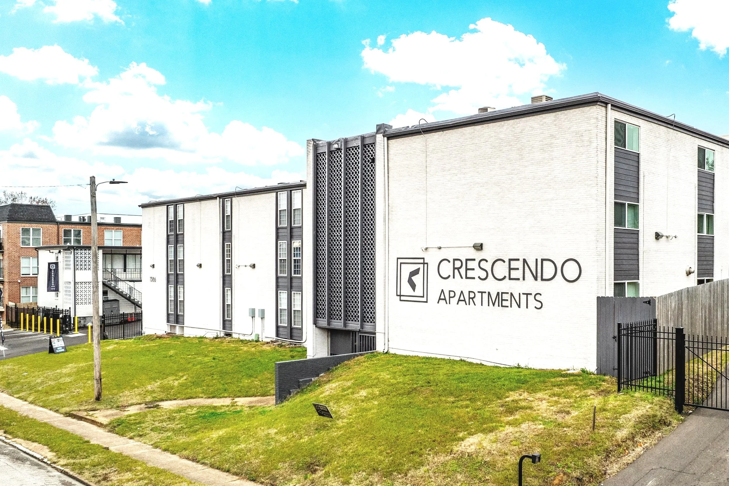 Exterior view of Crescendo Apartments, a modern multi-story apartment building with white and gray facade, black trim, and several windows, next to a grassy area and sidewalk under partly cloudy sky.