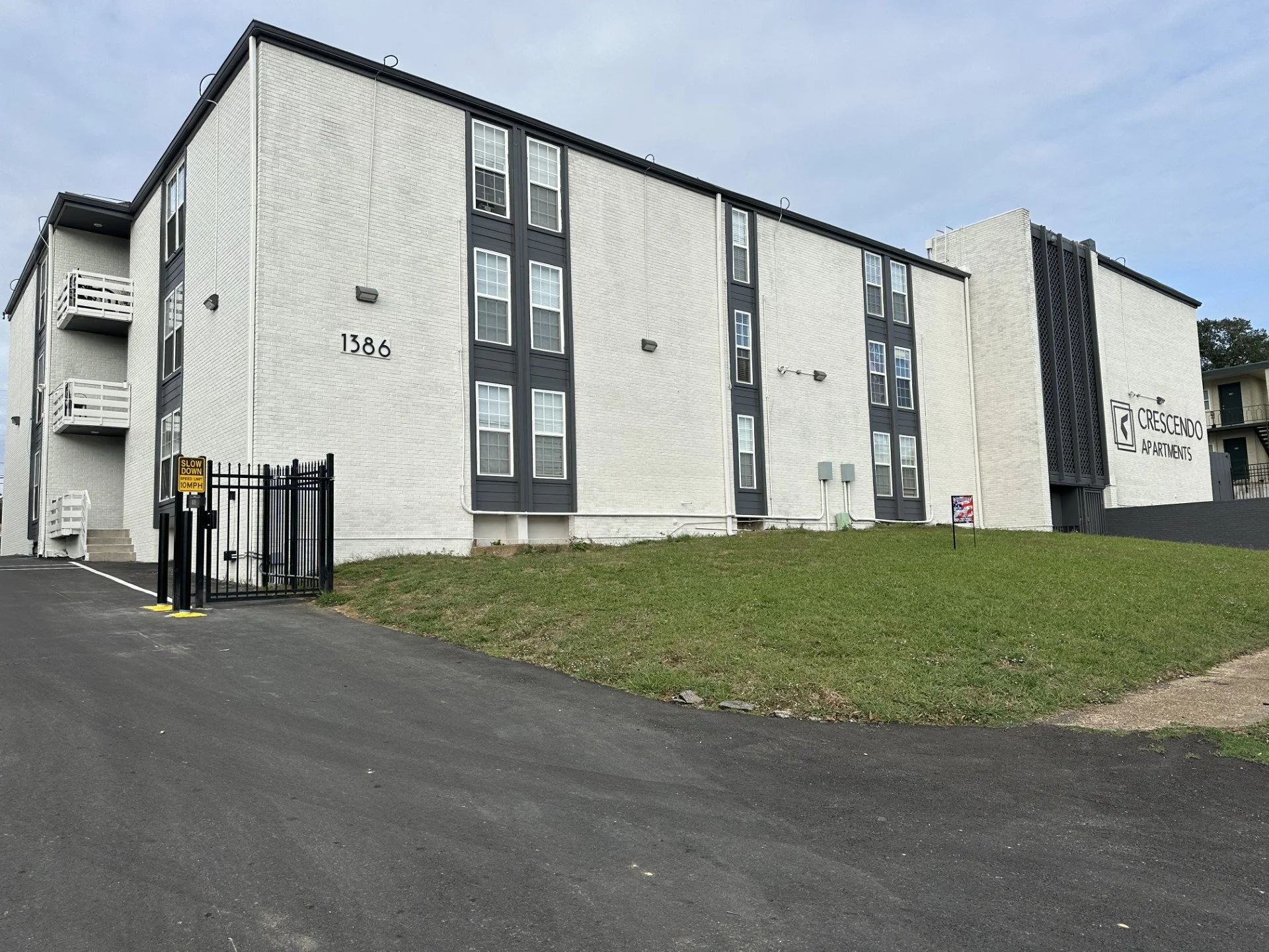 The exterior of a white apartment building with black accents and multiple windows. The building has a sign that reads 'Crescendo Apartments' and the number 1386. There is a black fence with a gated entrance and a small grassy area. The sky is cloudy