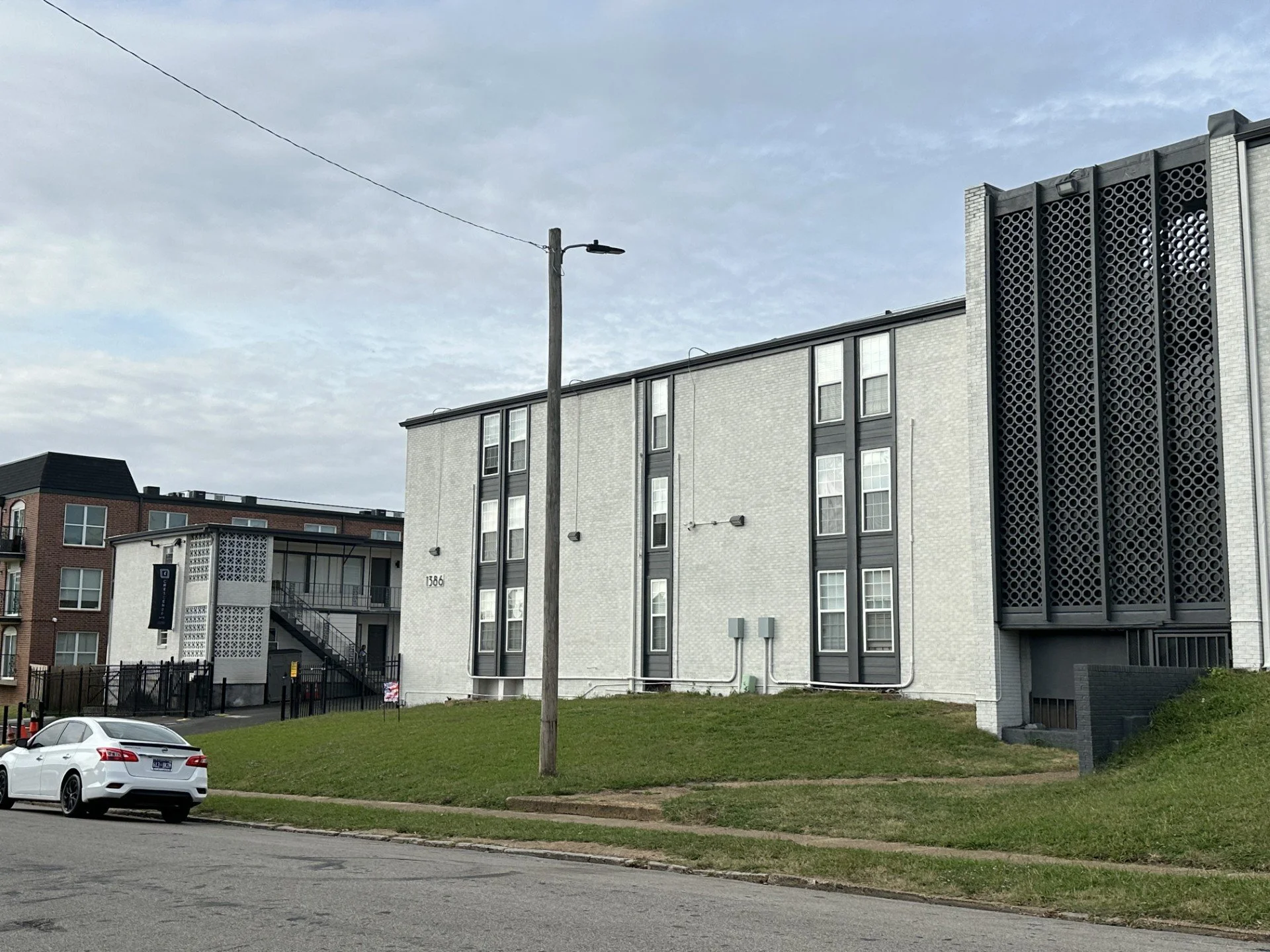 A modern multi-family residential building with a white brick exterior and black accents. There is a grassy area in front, a street with a white car parked, and a utility pole with an old-fashioned street lamp. The sky is partly cloudy.