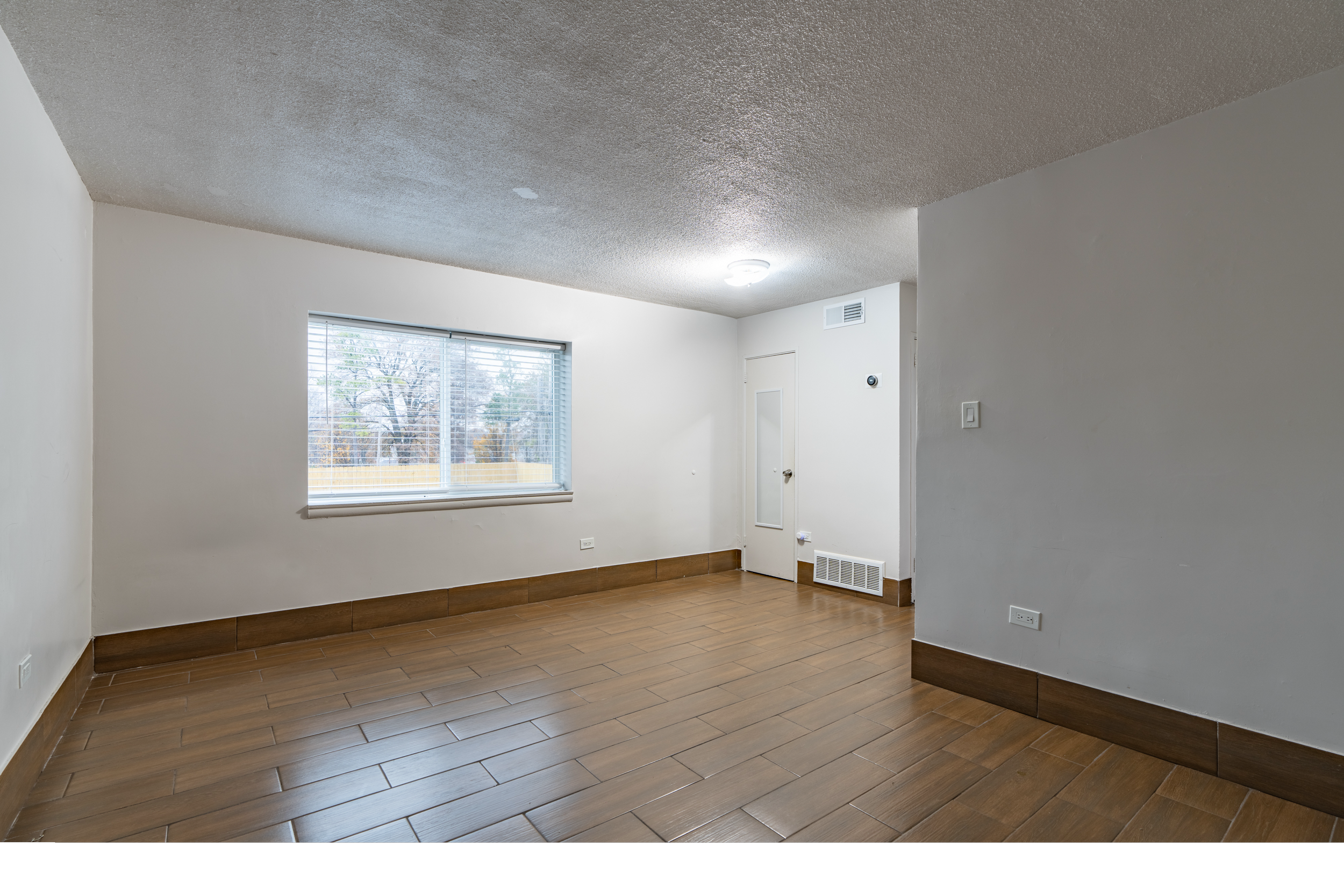 Empty living room with white walls, a large window with blinds, hardwood flooring, and ceiling light fixtures.