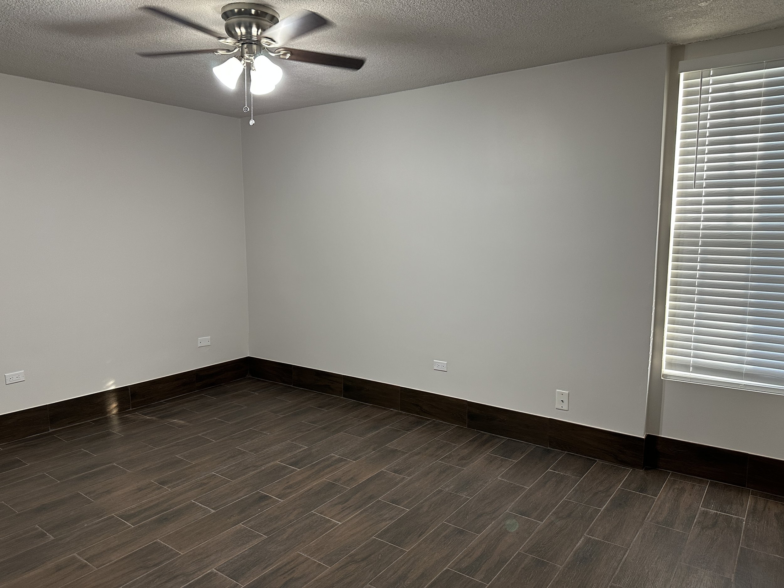 Empty room with a ceiling fan, white walls, dark tiled flooring, a window with blinds, and multiple electrical outlets.
