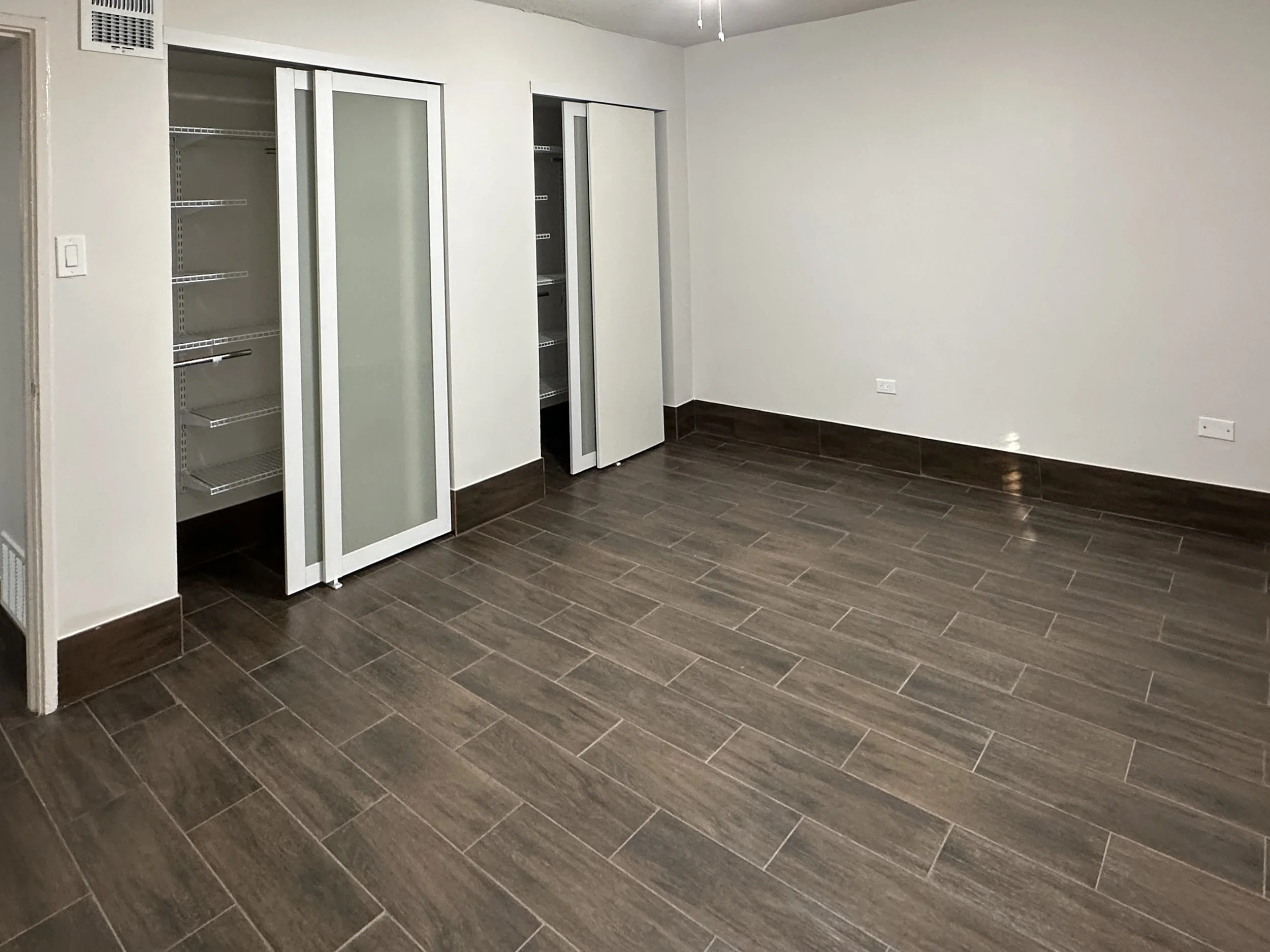 Empty room with white walls, dark tiled flooring, and built-in closets with sliding doors.