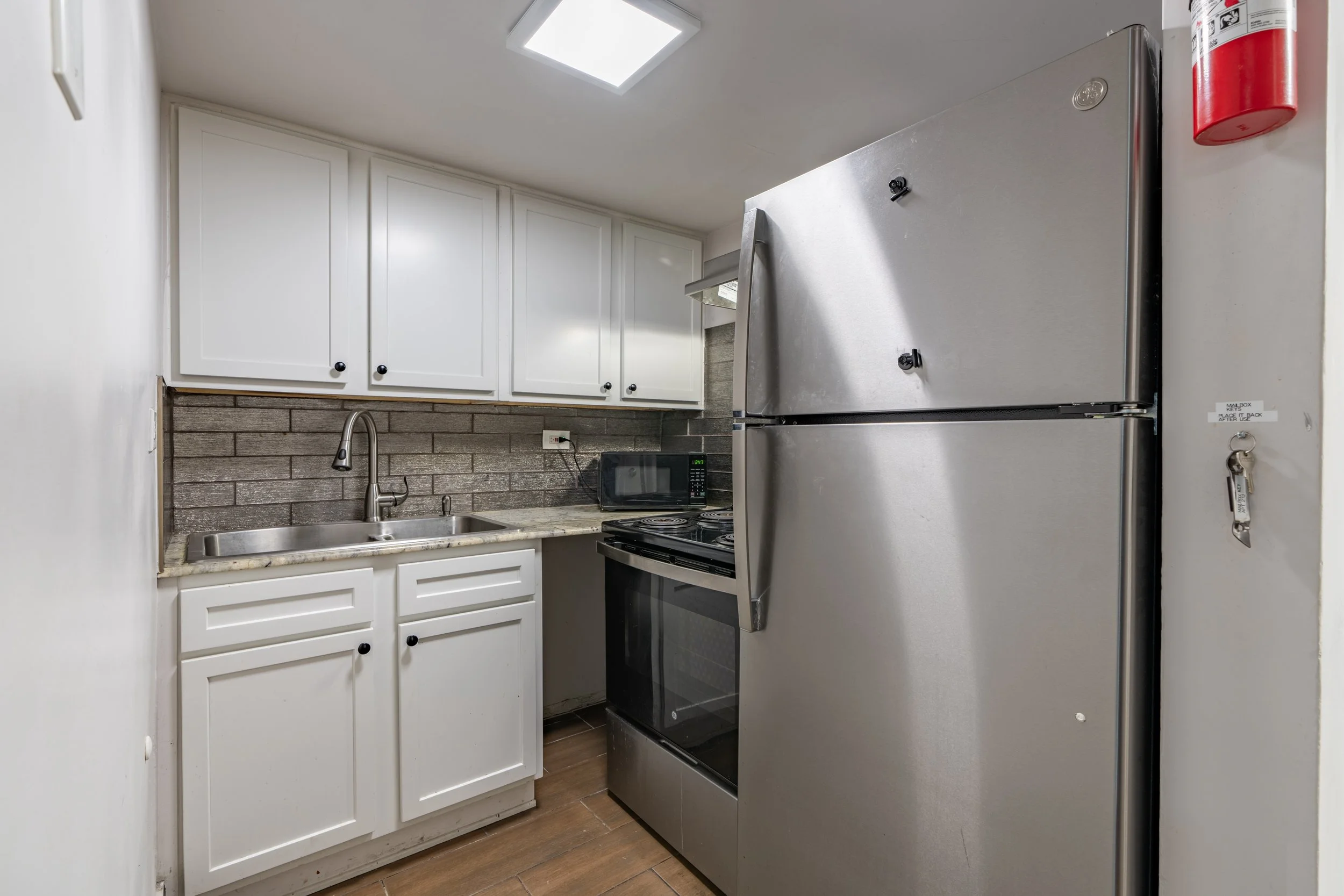 A compact kitchen with white cabinets, a stainless steel refrigerator, a small black microwave, a stove, and a gray brick backsplash. There is a sink with a silver faucet and a fire extinguisher mounted on the wall.