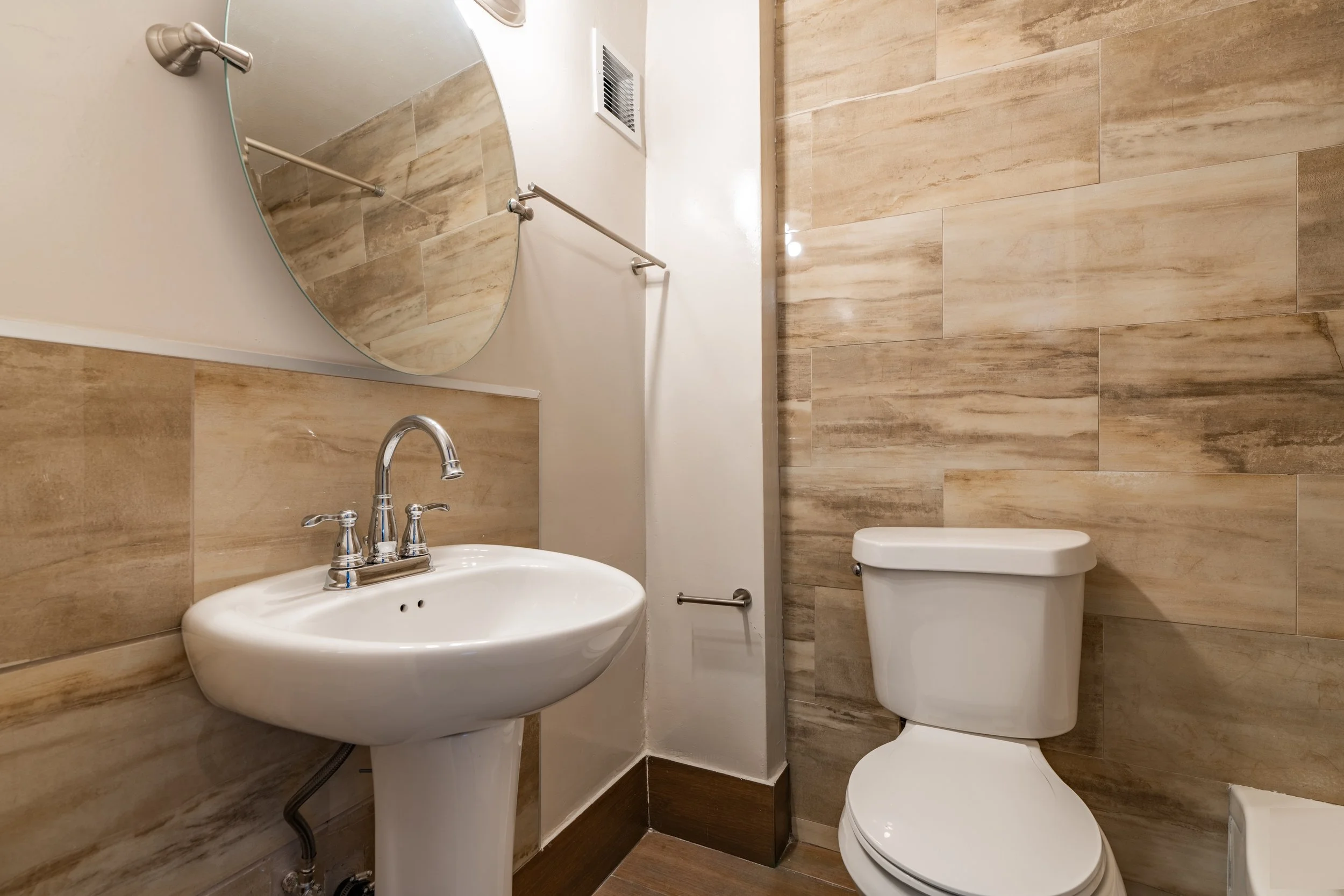 A small bathroom with a beige tiled wall, a white toilet, a white sink with chrome fixtures, a large mirror, and a wall vent. There is a metal handrail above the toilet and a curved metal pipe connected to the sink.