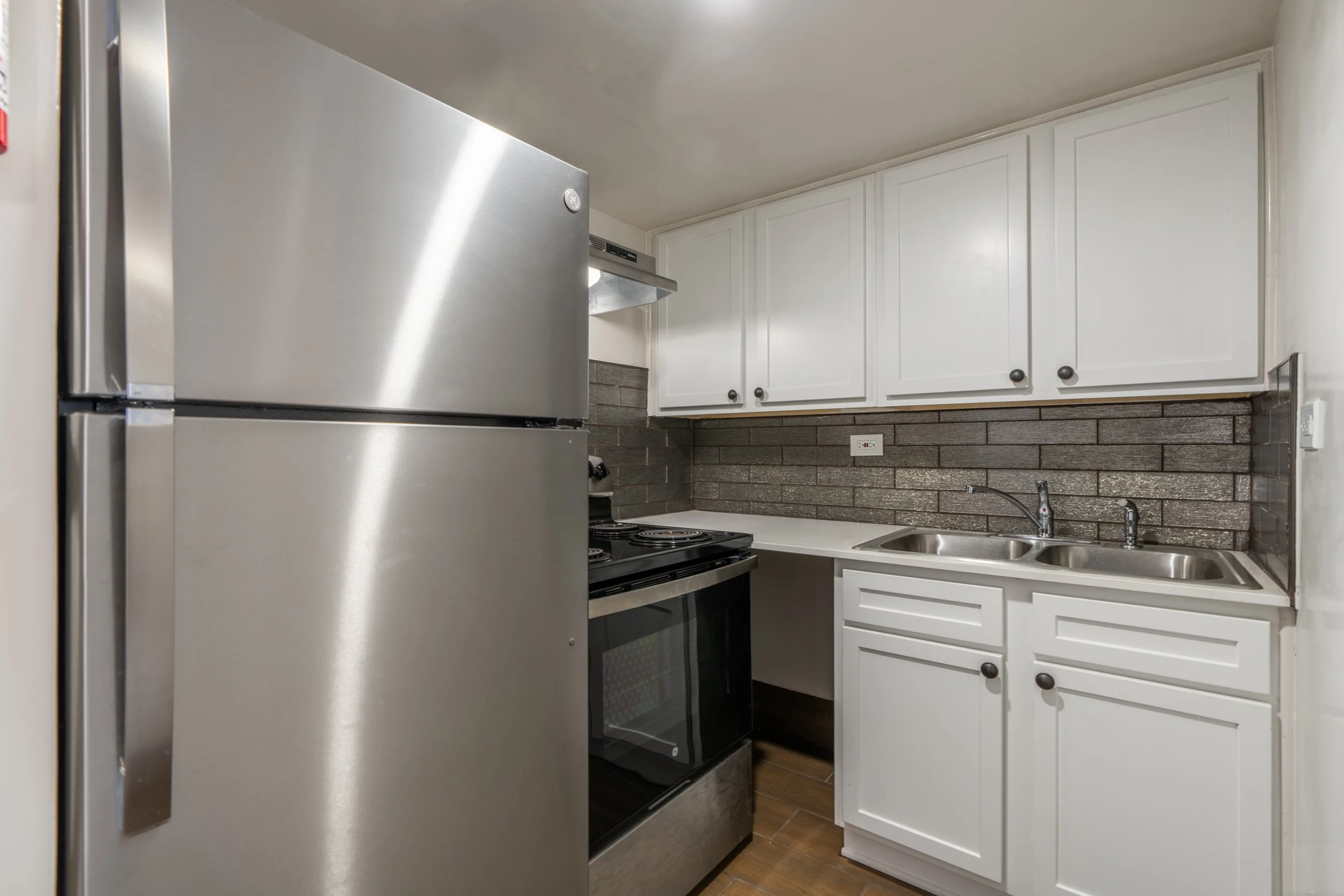 Small kitchen with white cabinets, gray tile backsplash, stainless steel refrigerator, stove, and double sink.