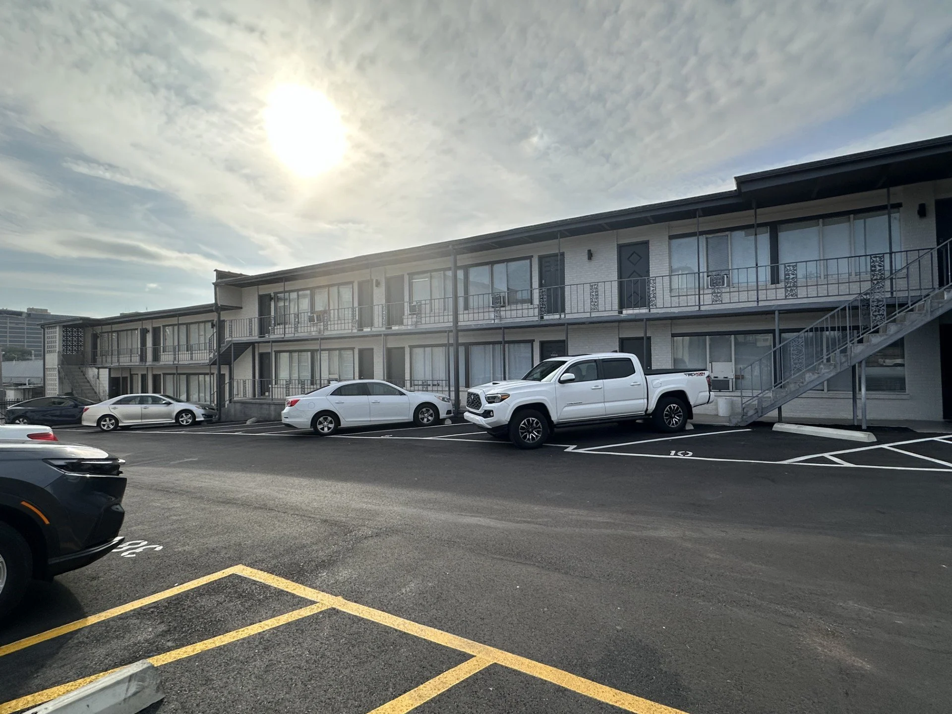 Multi-story apartment complex with parking lot in front, several cars parked, and a partly cloudy sky with the sun shining.