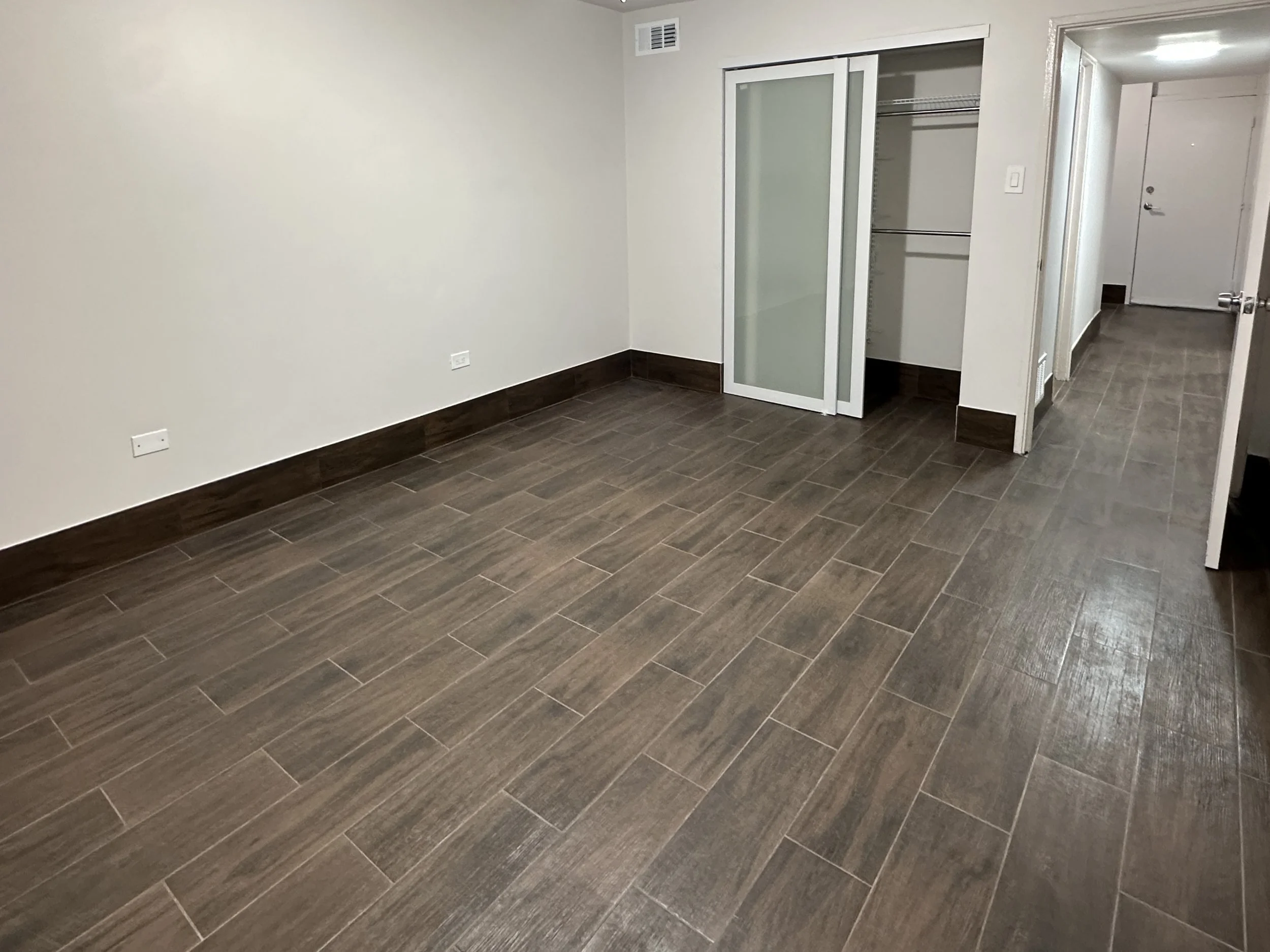 Empty room with dark wooden floors, white walls,wardrobe with frosted glass sliding door, open closet with metal rod, and the entrance door in the background.