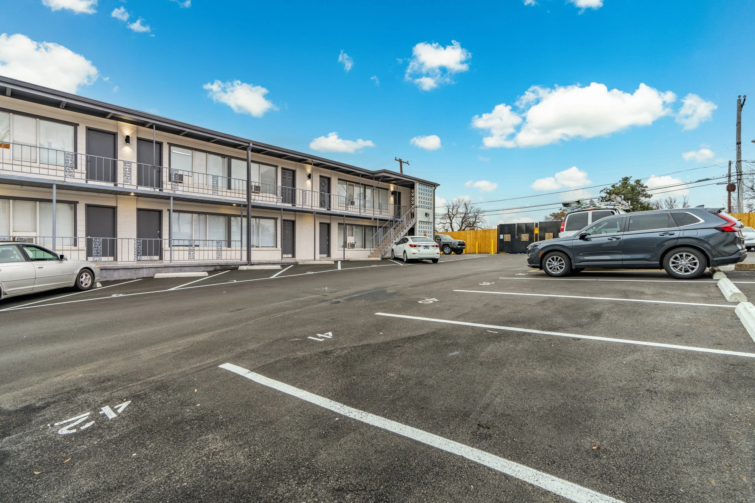 Parking lot outside a two-story apartment building under a blue sky with scattered clouds. Several cars are parked in the lot, including a white sedan, a black SUV, and a gray crossover. The building has black doors and doors with large windows, with