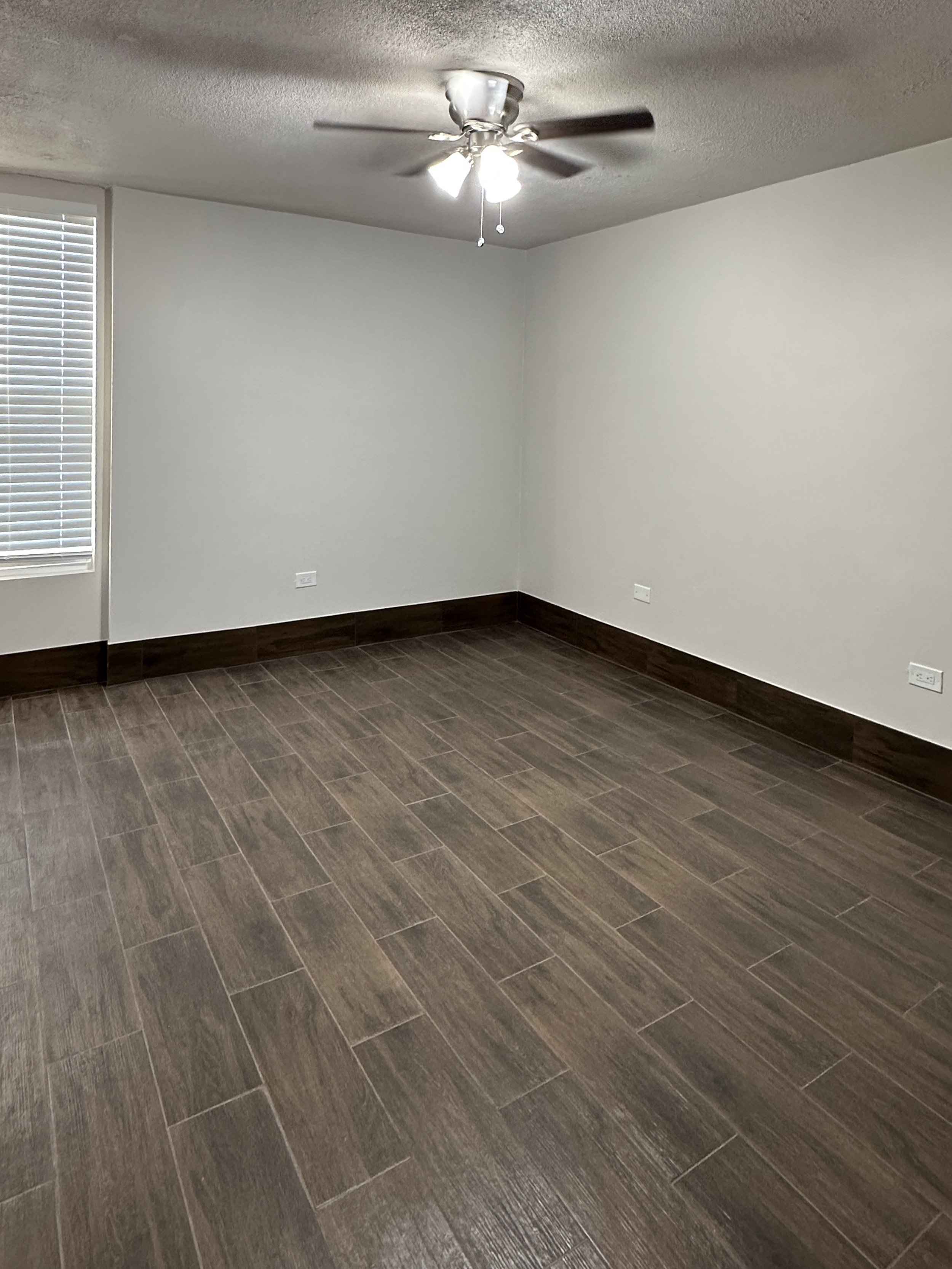 Empty room with brown wooden tile flooring, white painted walls, a window with closed blinds, and a ceiling fan with light.