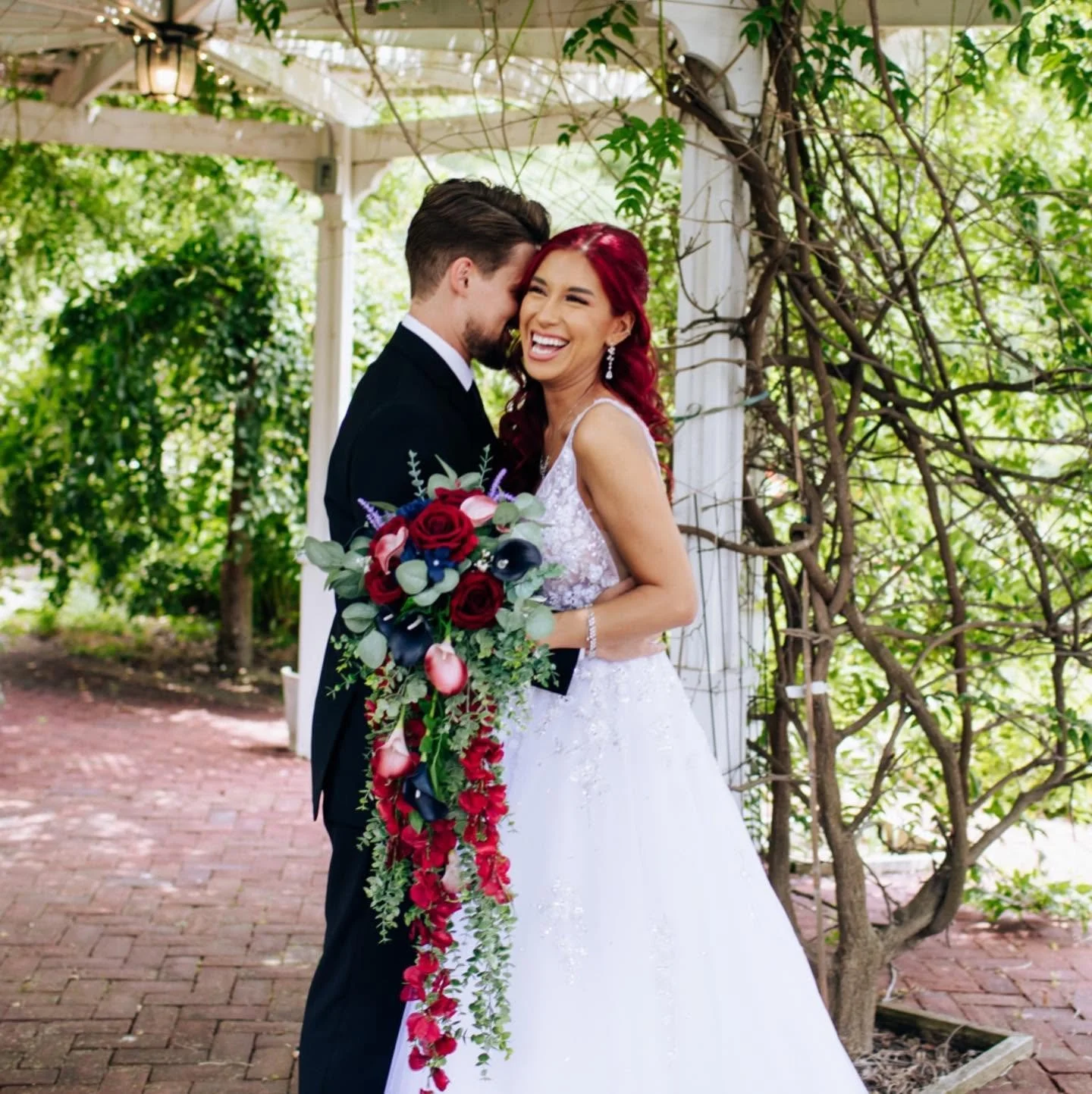 A bride and groom on their wedding day, standing close together outdoors. The bride has red hair and is wearing a white wedding gown, holding a long bouquet of red, pink, and dark blue flowers. They are smiling and sharing a moment of happiness.