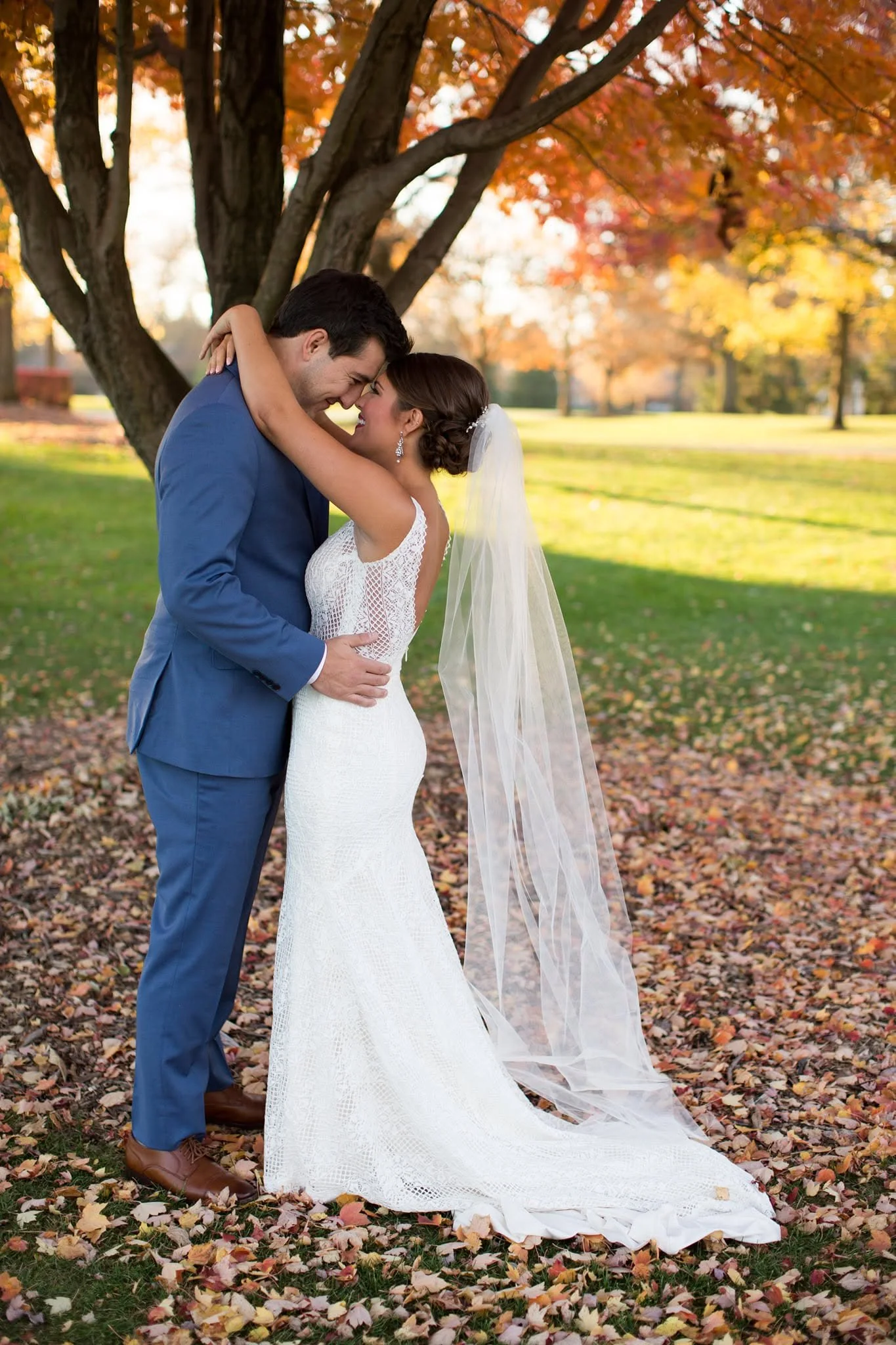 A newlywed couple in formal wedding attire standing near a large tree in a park during autumn. They are smiling, embracing, and touching foreheads amidst fallen leaves with vibrant fall foliage in the background.