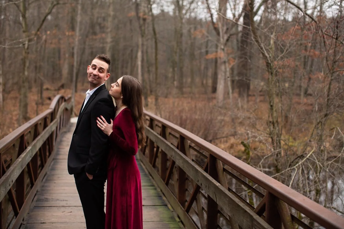 A couple in formal attire standing on a wooden bridge in a forest during autumn, with leafless trees and fallen leaves in the background.