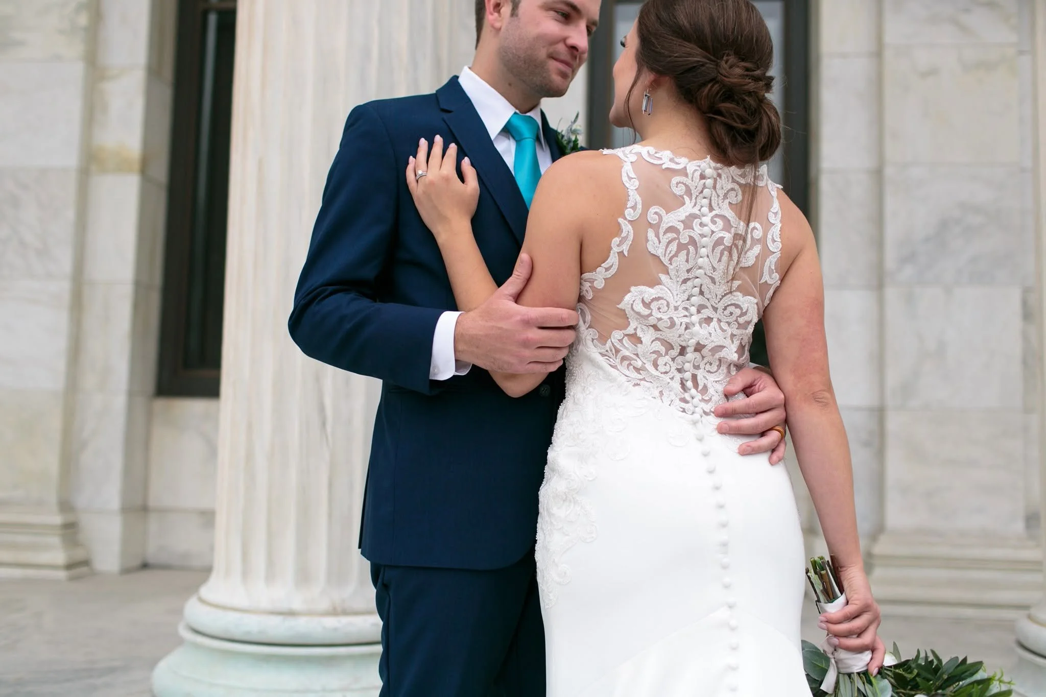 A bride and groom hold each other closely during their wedding, standing outdoors in front of white columns. The groom is dressed in a navy blue suit with a white shirt and turquoise tie, while the bride wears a white lace wedding gown with an open back and intricate floral embroidery. The bride has her hair styled in an elegant updo and is wearing earrings, and she holds a small bouquet of flowers in her right hand.