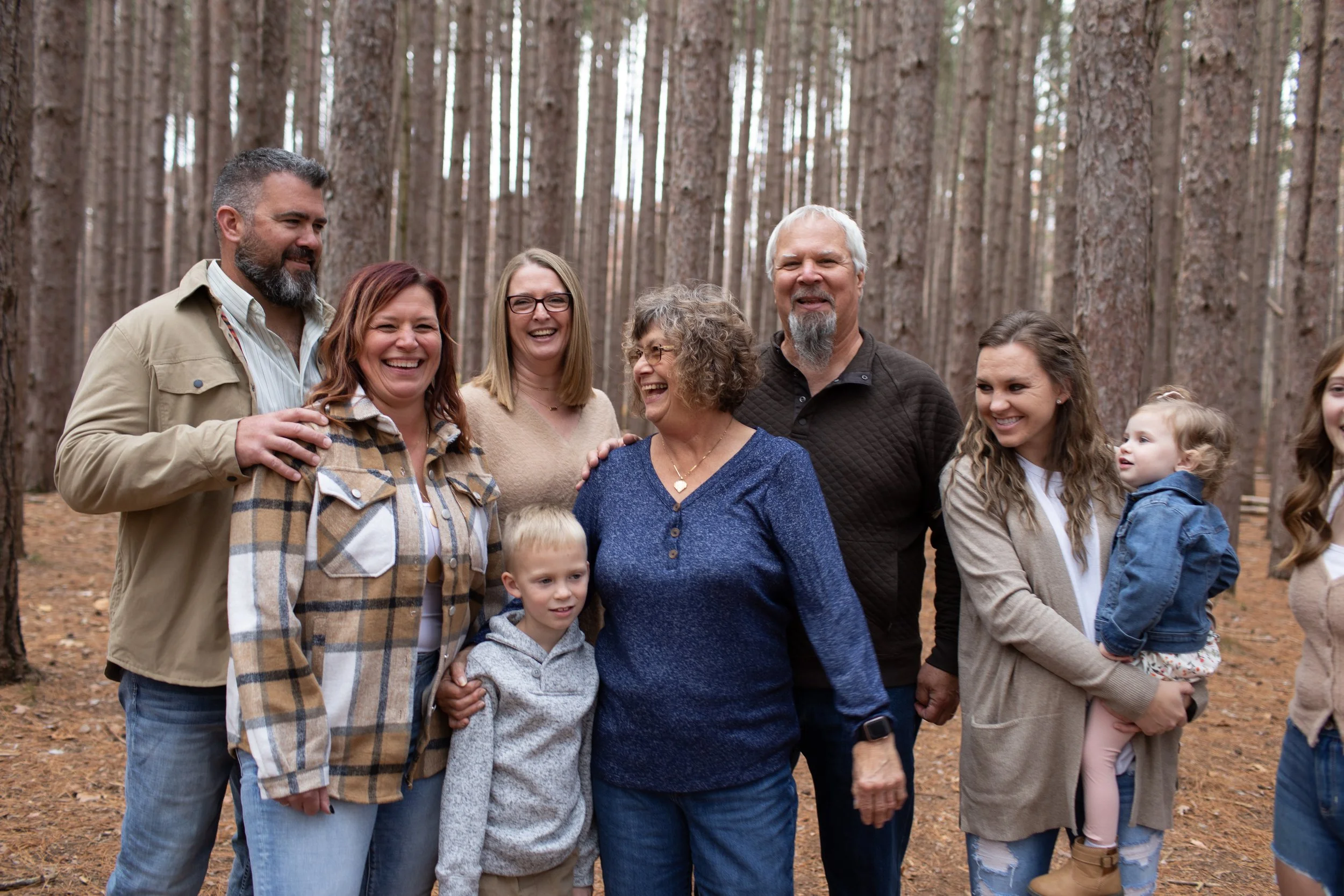 Group of people standing outdoors in a pine forest, smiling and talking, with children and adults of various ages.