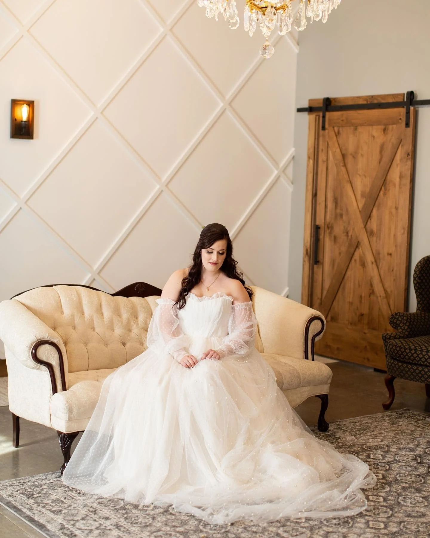 A woman in a white wedding dress sitting on a vintage cream-colored sofa in a room with a rustic wooden sliding barn door, a chandelier overhead, and a beige and cream patterned rug on the floor.