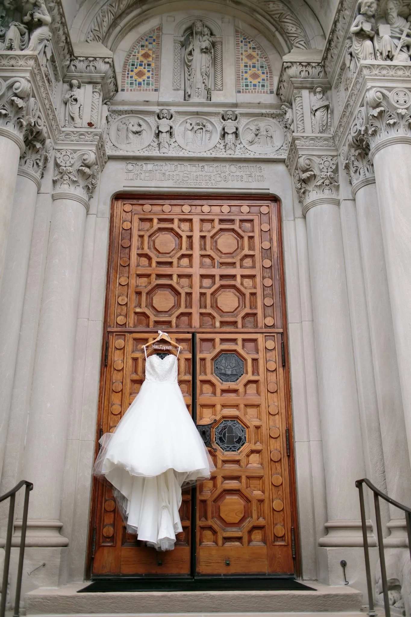 A white wedding dress hanging on a hook in front of a grand, ornate wooden door with intricate carvings, set within an elaborate stone archway with sculpted figures and decorative elements.