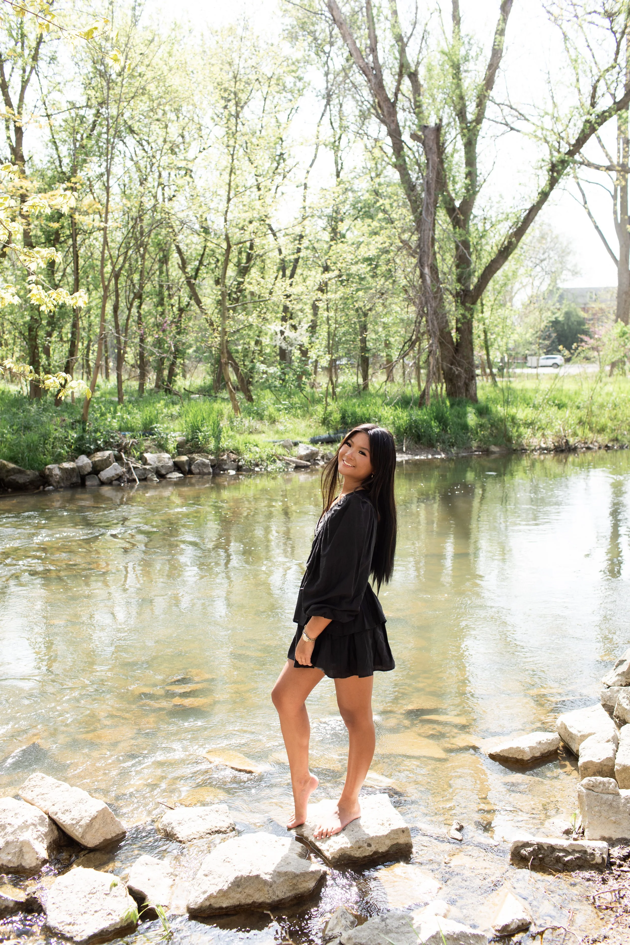 A young woman standing on rocks in a shallow river with trees and greenery in the background on a sunny day.