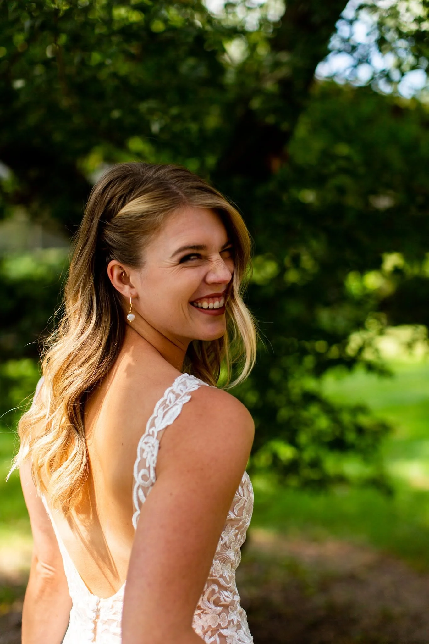 A woman with long wavy hair smiling outdoors, wearing a white lace dress with orchid earrings, in front of green trees.