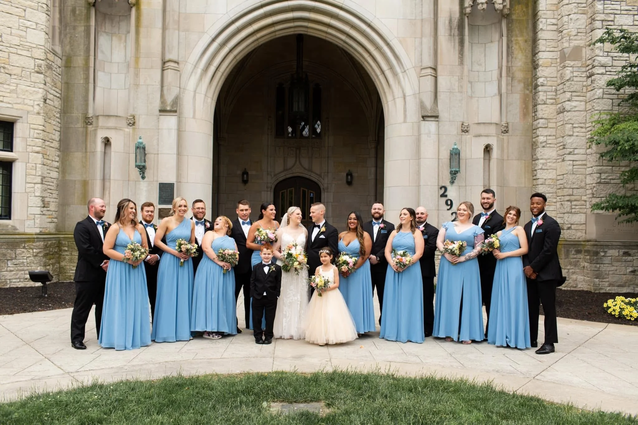 A large wedding party posed outside a stone church building, with the bride and groom in the center, surrounded by bridesmaids and groomsmen, all dressed in formal attire.