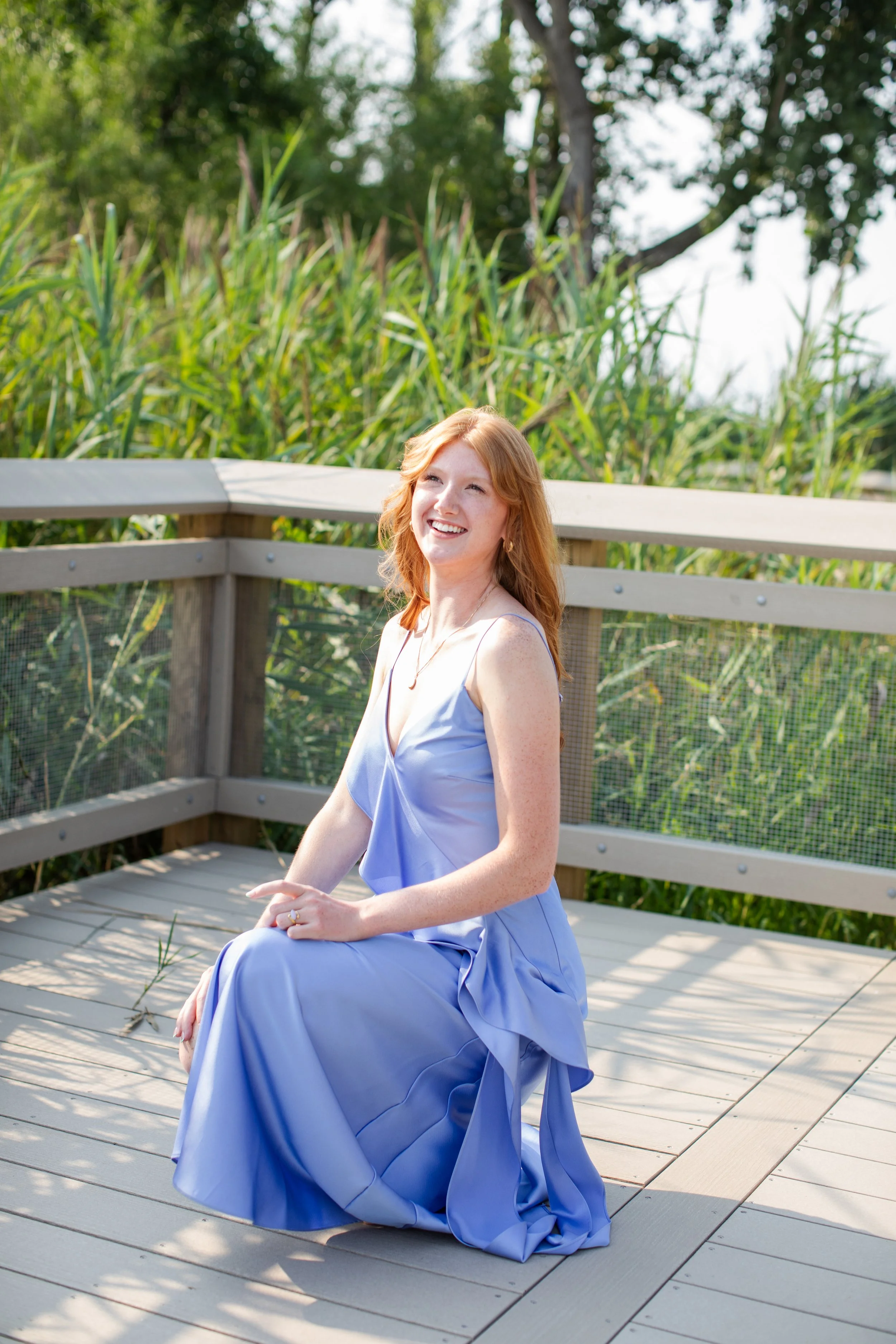 A woman with red hair and a light purple dress sitting on a wooden deck, smiling, with green trees and tall grasses in the background.