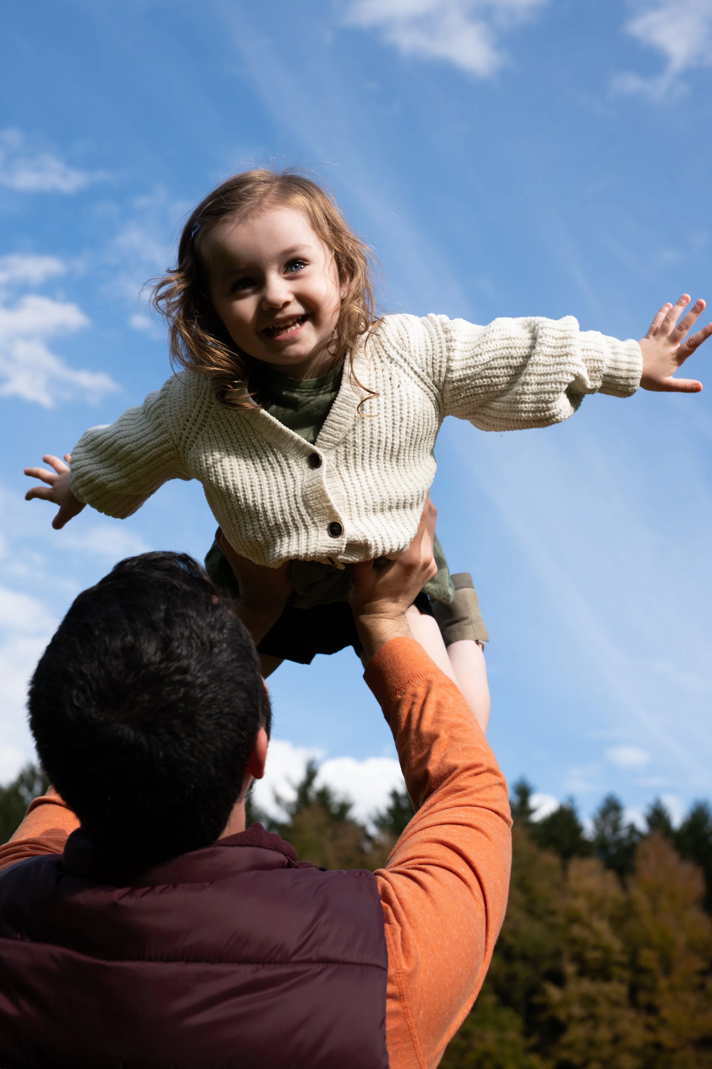 A young girl with curly hair smiling, arms outstretched, sitting on an adult's shoulders outdoors with a clear blue sky and trees in the background.