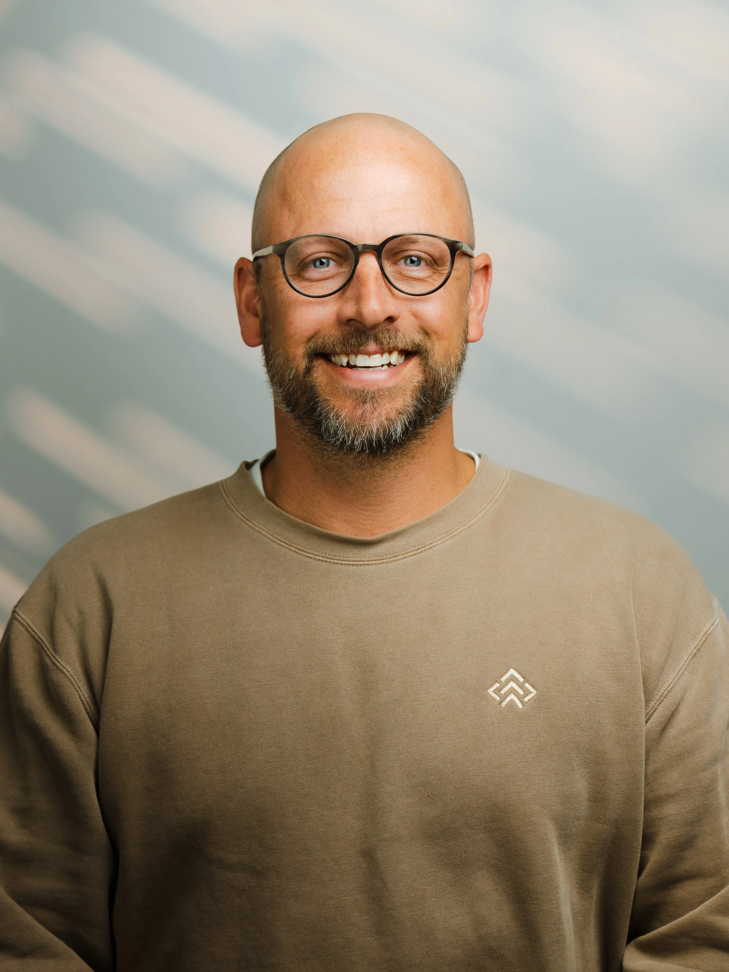 A smiling man with glasses, a beard, and a bald head wearing a beige sweatshirt with a small logo on the chest.