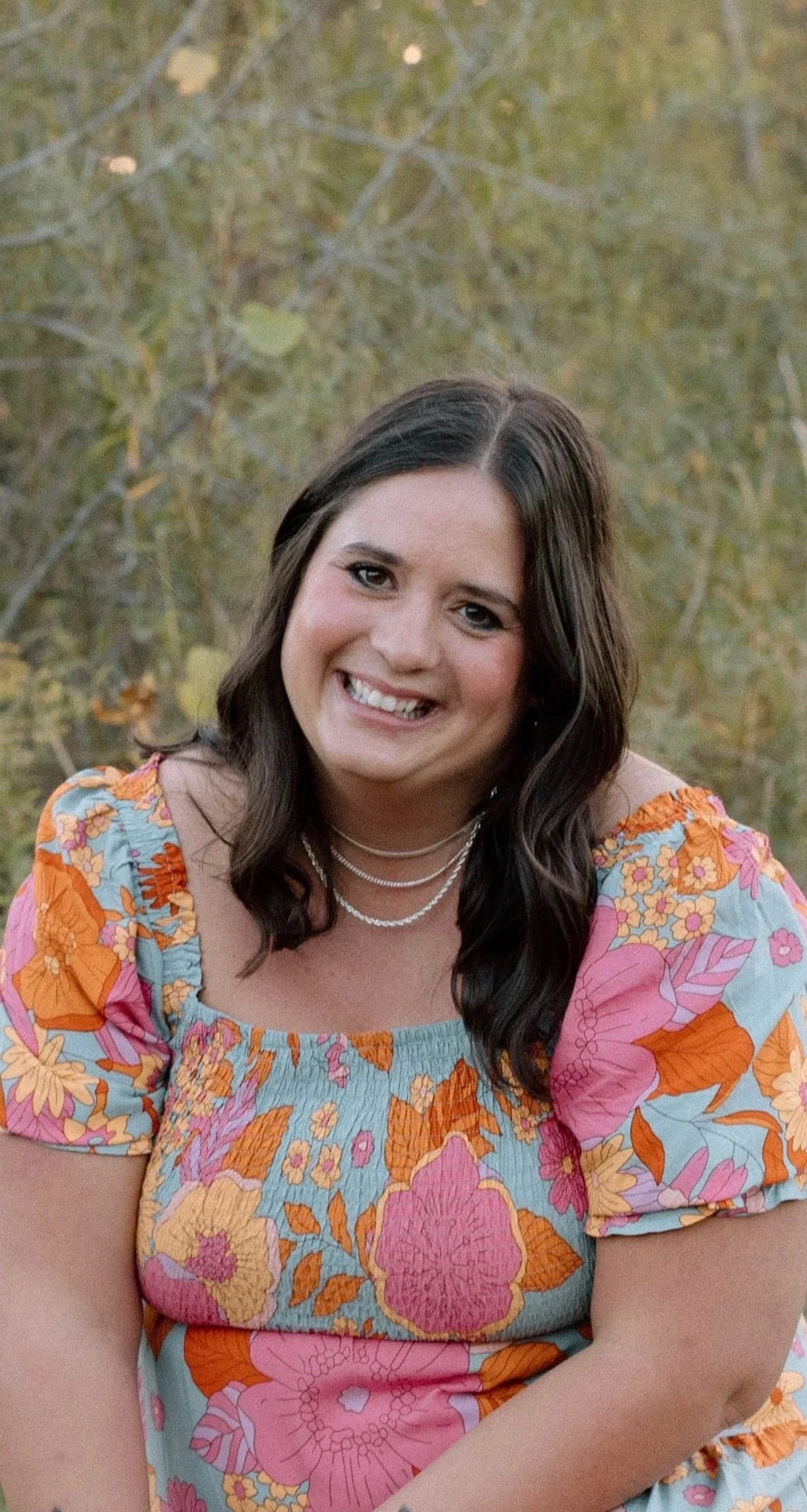 A woman with dark wavy hair smiling, wearing a colorful floral dress with orange, pink, and blue flowers, outdoor background with trees.