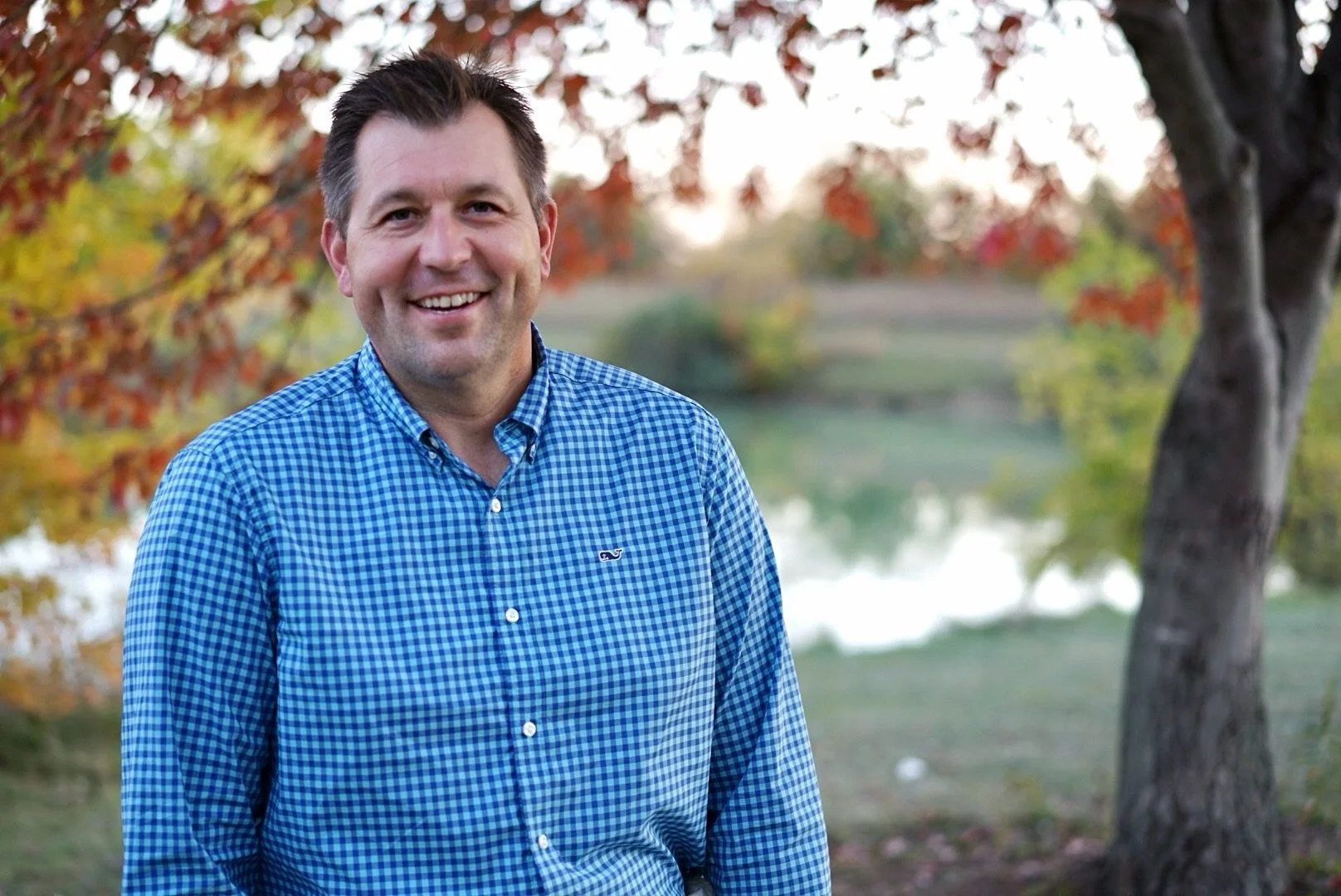 A smiling man wearing a blue checkered shirt standing outdoors near a tree with autumn-colored leaves, a lake, and a pathway in the background.