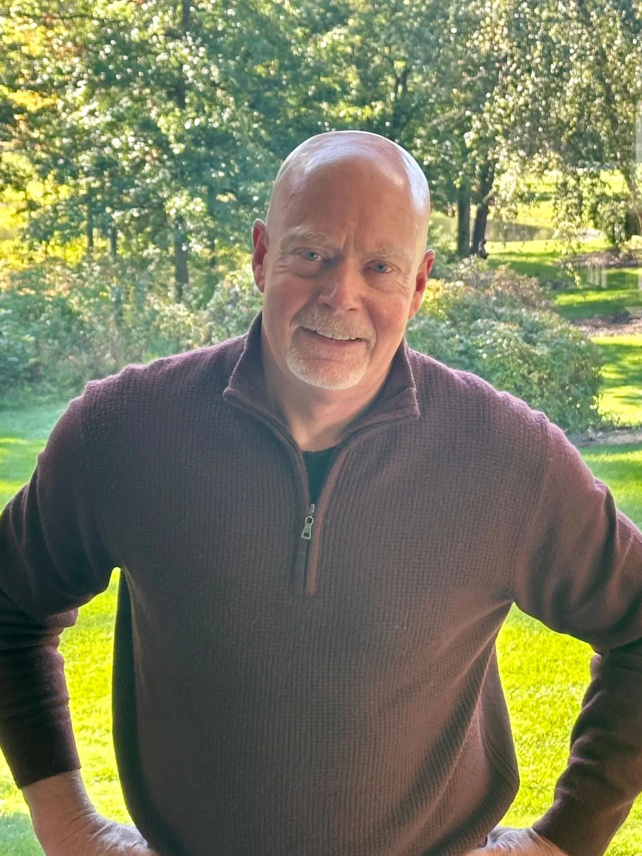 A middle-aged man with a bald head and light facial hair smiling outdoors in front of green trees and grass.