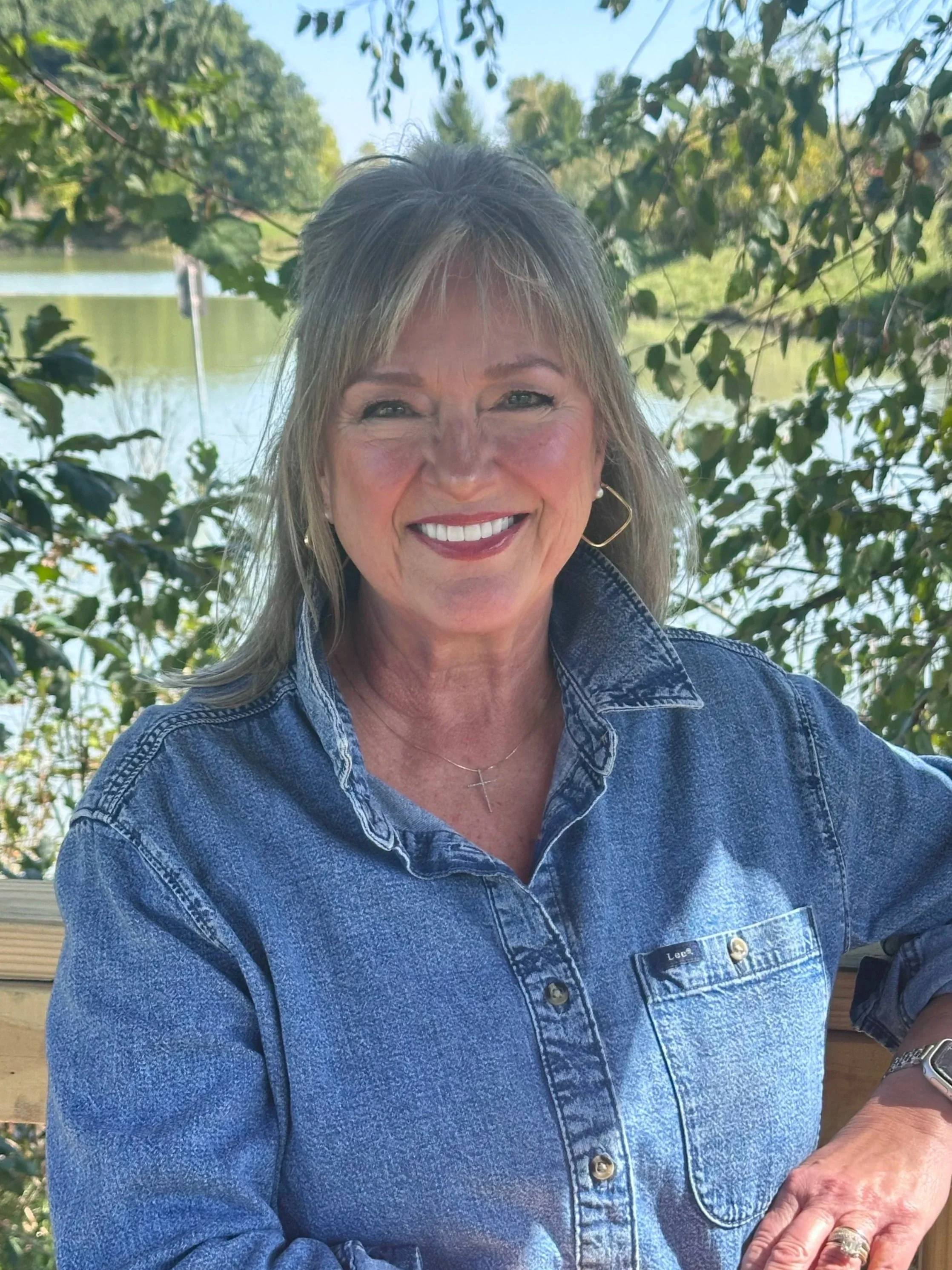 A smiling woman with blonde hair and hoop earrings wearing a blue denim jacket, standing outdoors with trees and a lake in the background.