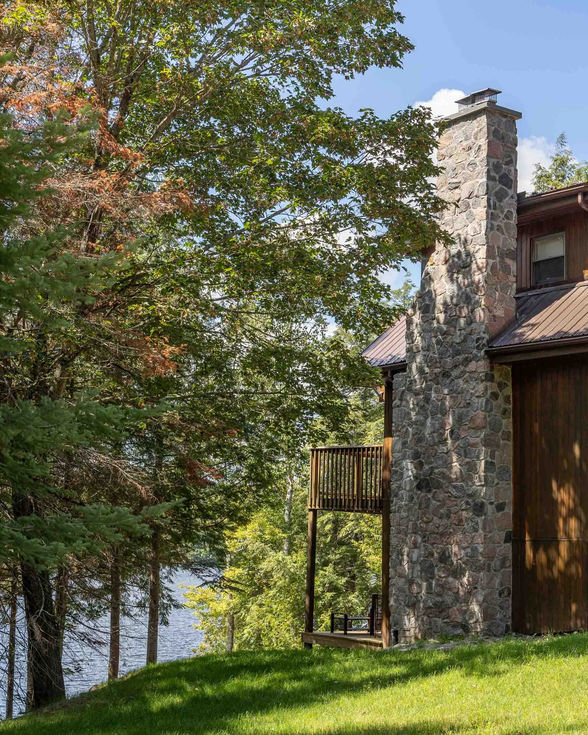 The image shows a house with a stone exterior and a chimney, surrounded by trees with green leaves, and a body of water in the background. There is a small wooden deck with a railing attached to the house, and a grassy yard in the foreground.
