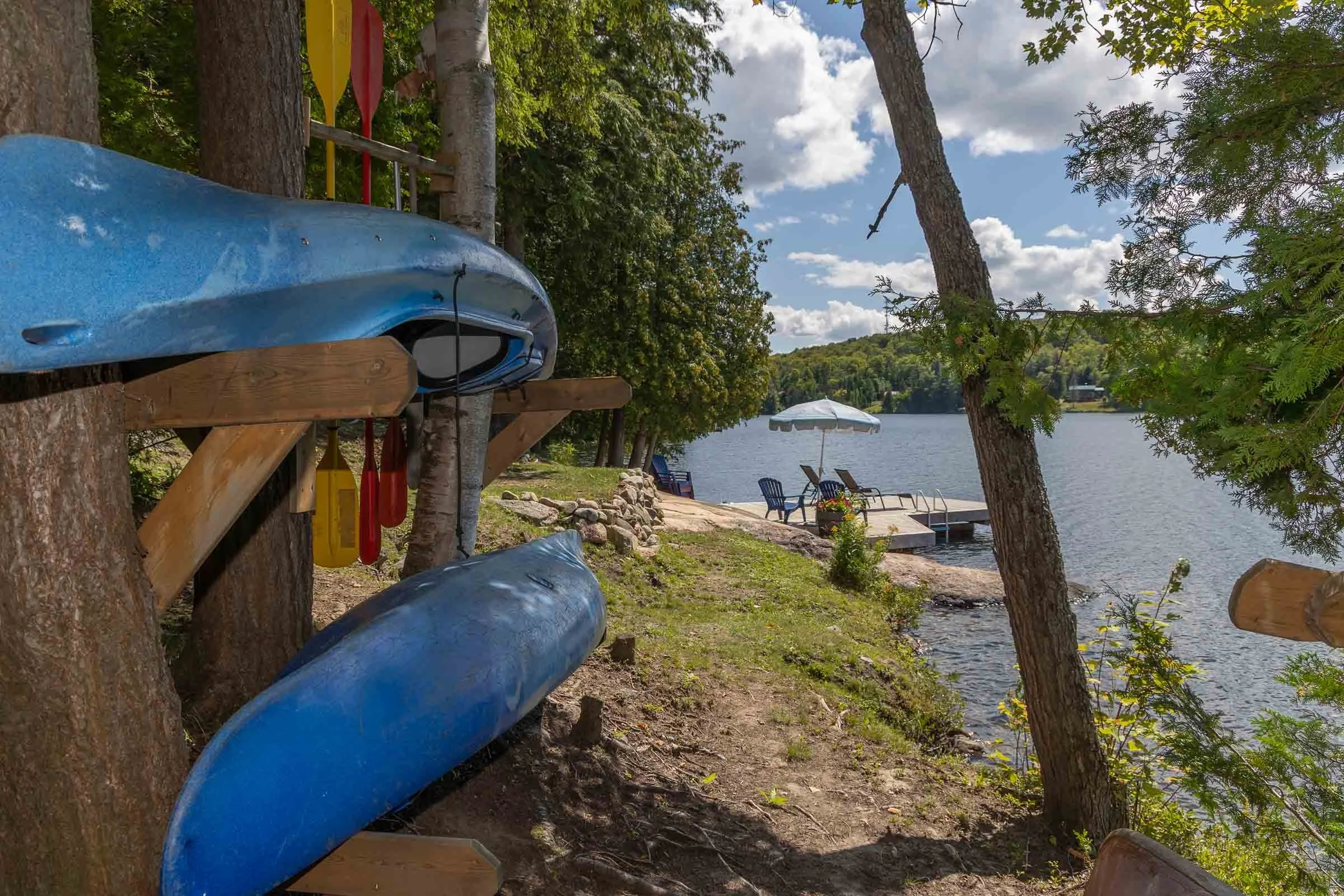 Blue canoes stored in a rack among trees near a lakeside with chairs, an umbrella, and a dock in the background.