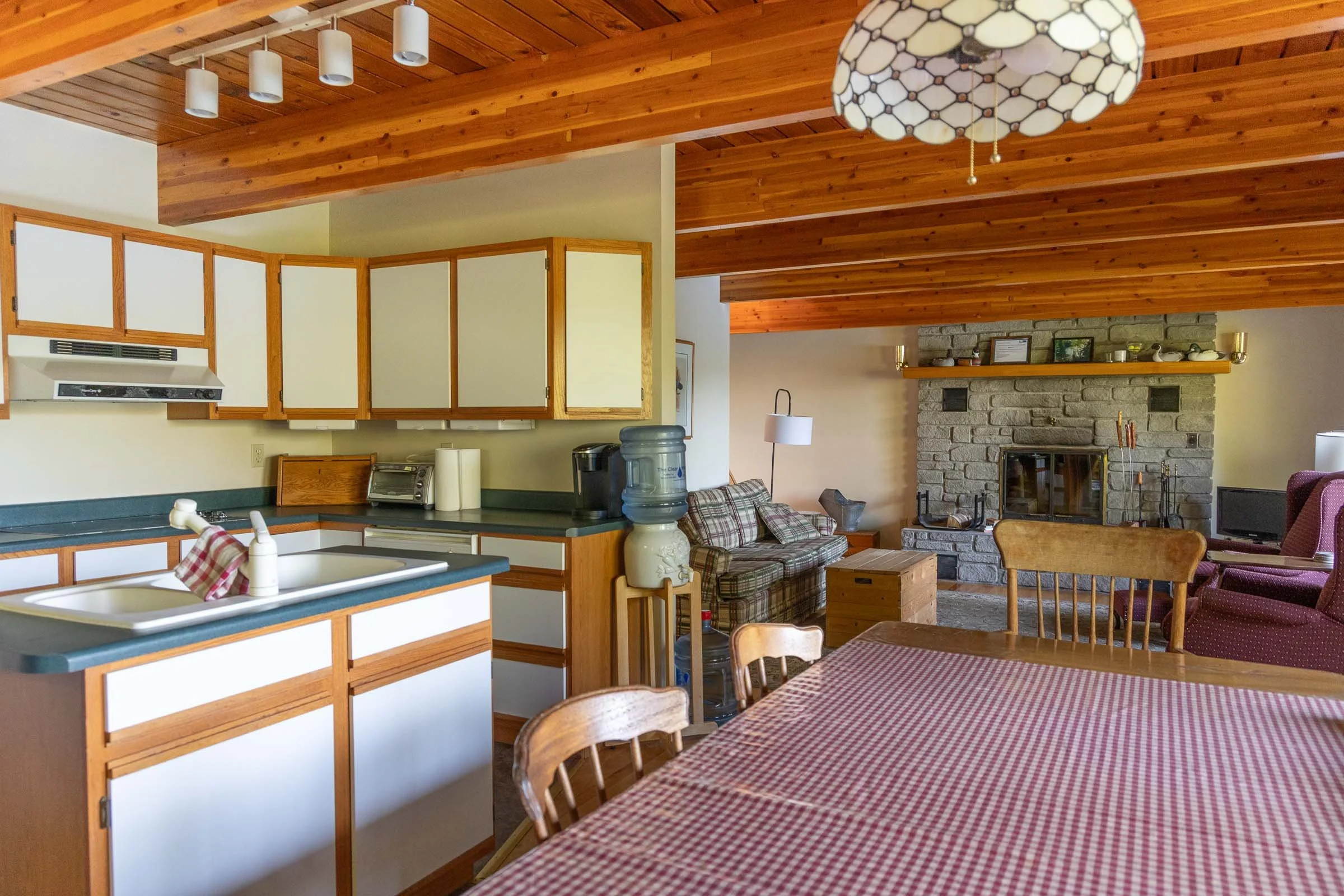 Interior view of a cozy living space with a wooden ceiling, a stone fireplace, and a kitchen area featuring white and wood cabinets, a water cooler, and a double sink with a checkered dish towel. There are also wooden dining chairs and a table with a